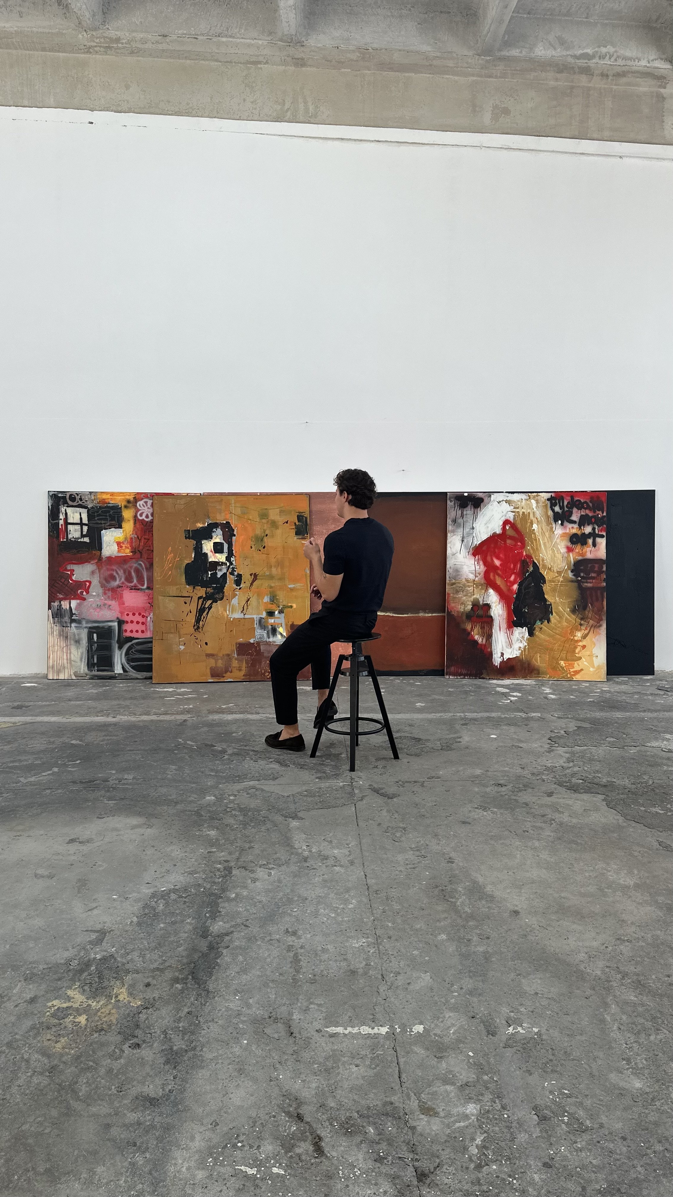Artist sitting on a stool in front of three abstract paintings displayed against a white wall in an industrial-style gallery space.