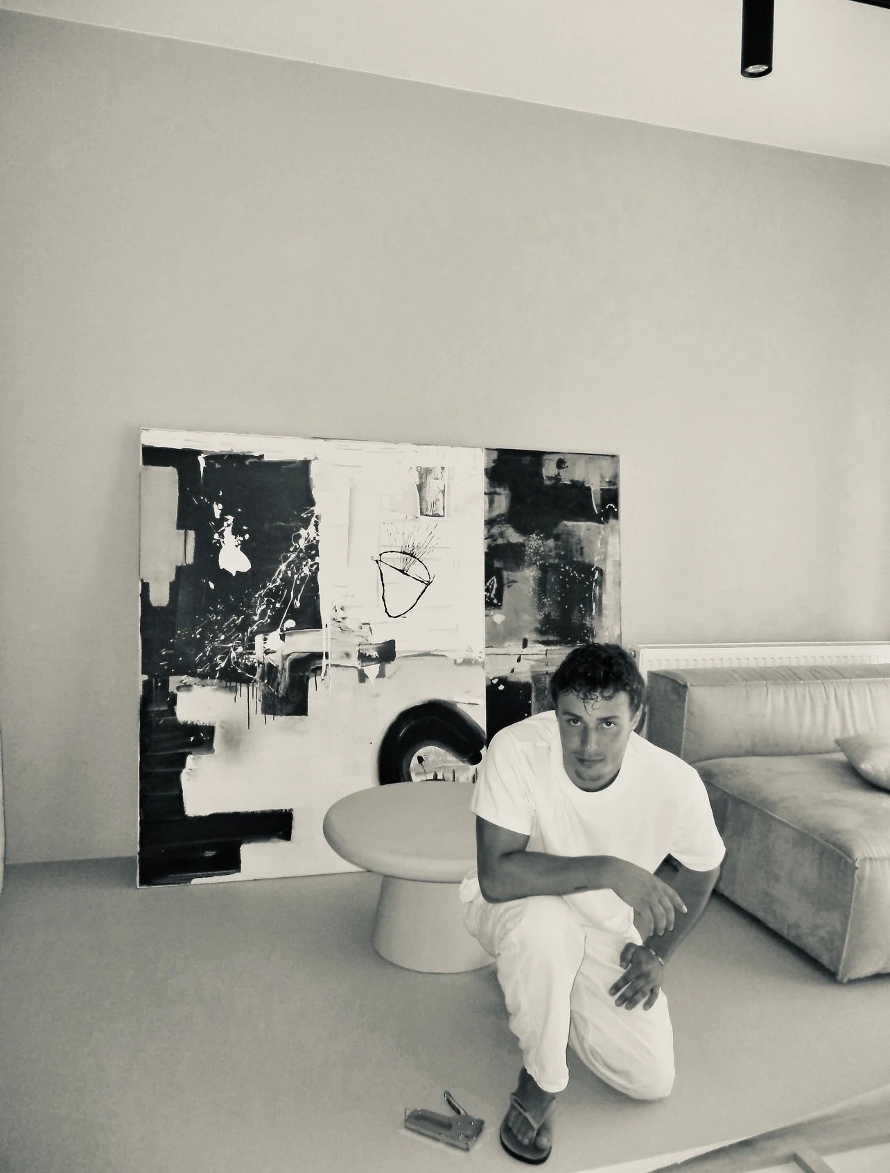 A young man with dark curly hair crouching in a living room with a large abstract black and white painting behind him, a beige sofa to his right, and a small round table in front of the painting.