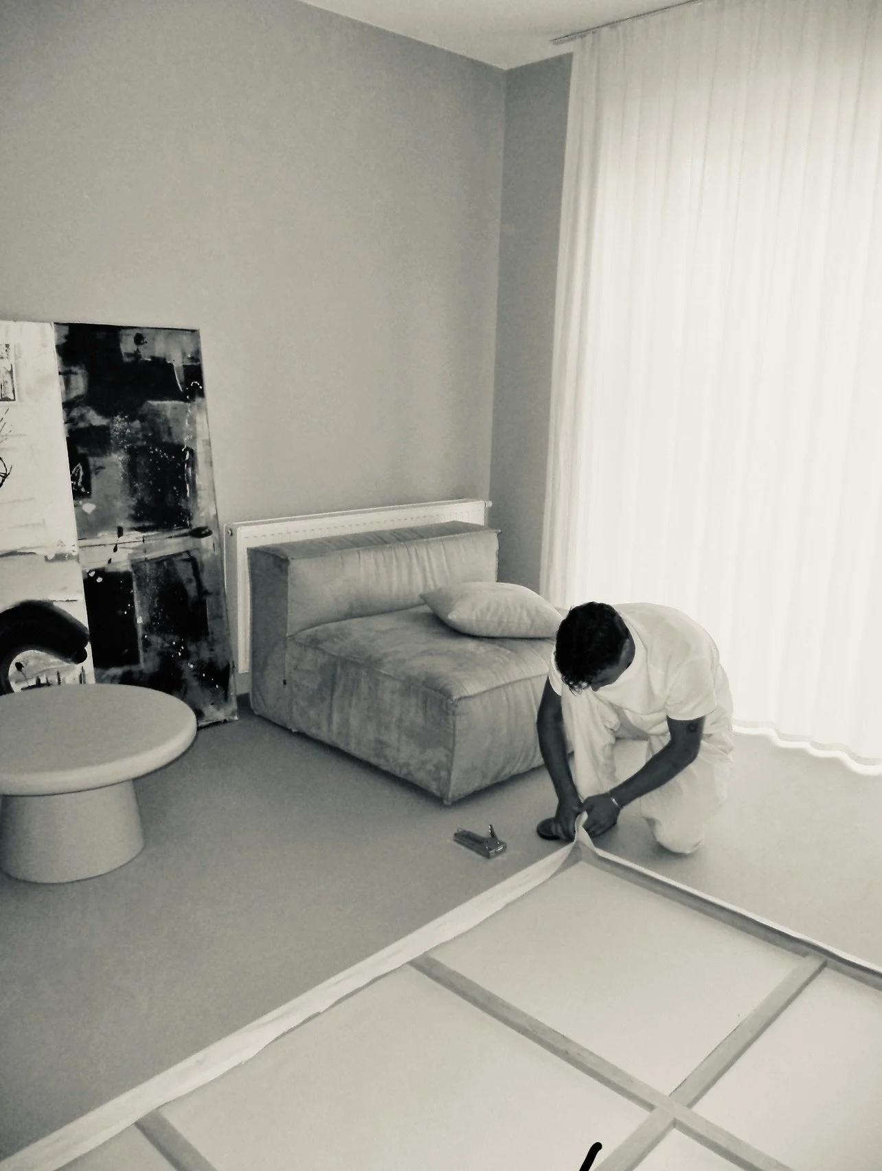 A person kneeling on the floor assembling a bed frame in a room with a gray sofa, a painting, and large white curtains