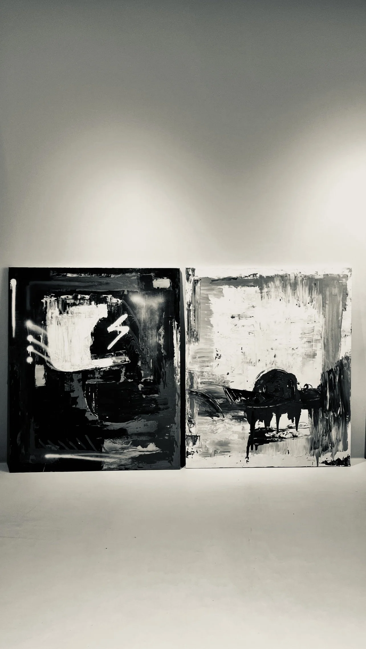 Two abstract black and white paintings on display, one featuring bold brushstrokes, a lightning bolt, and a silhouette of a face, the other depicting a figure with wings against a textured background.