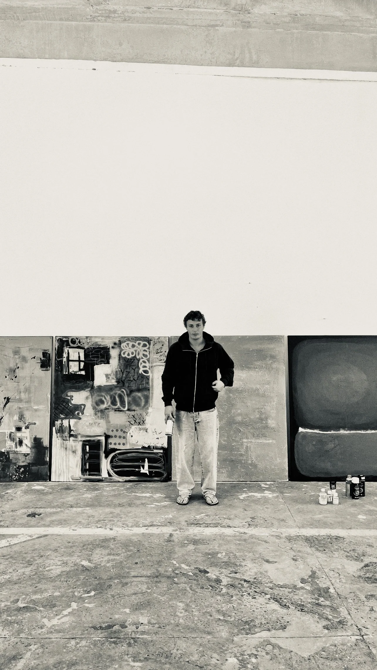A person standing in front of a large blank wall with paintings and artistic supplies on the ground nearby.