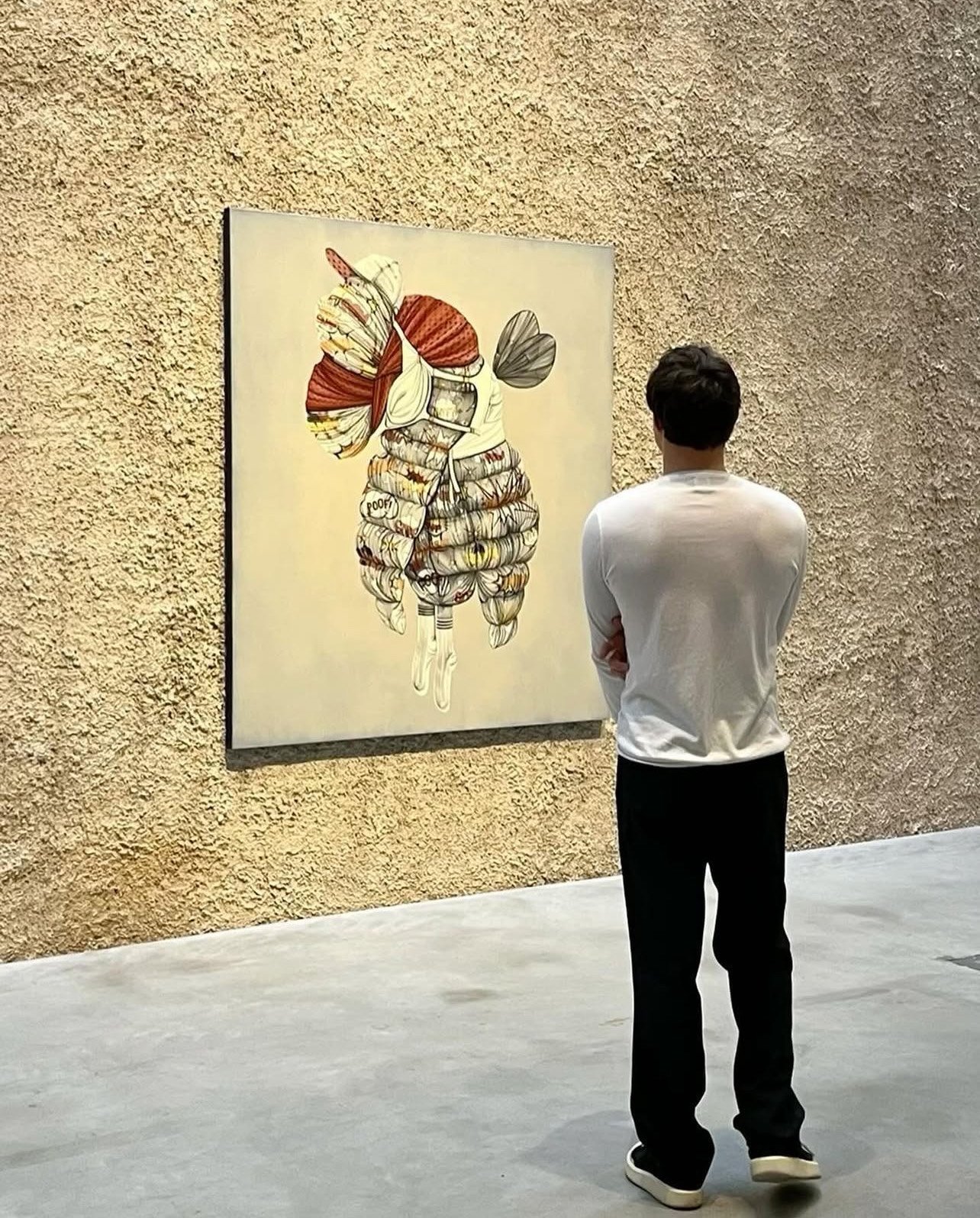 A man in a white shirt and black pants viewing an abstract art piece of a butterfly on a gallery wall.
