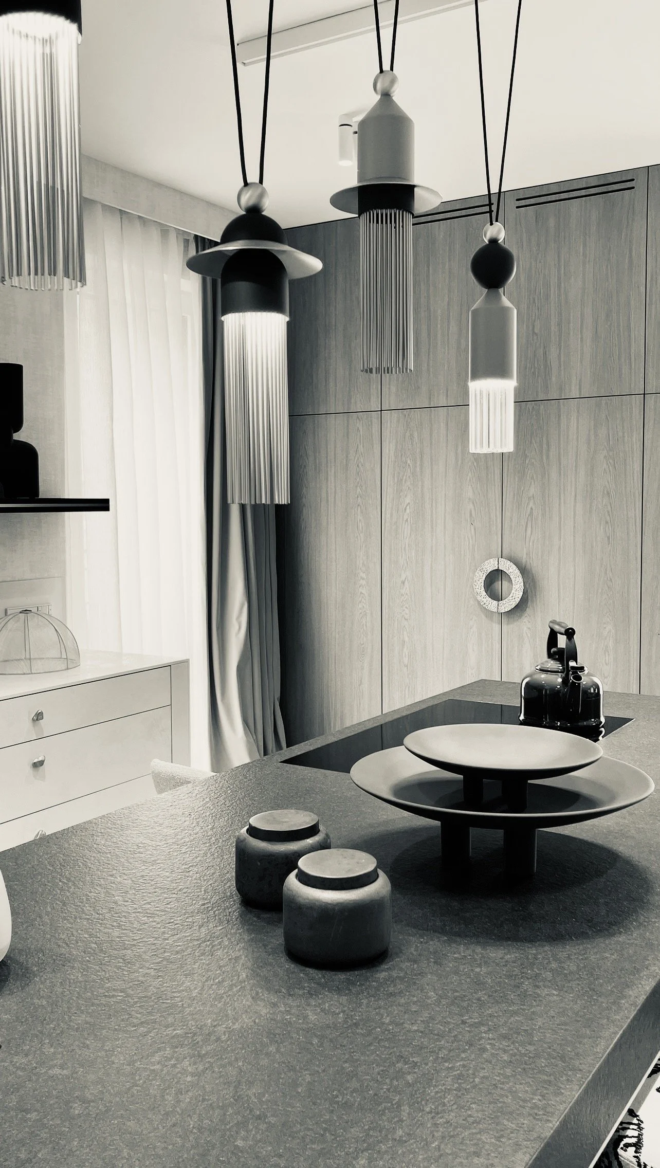 A modern kitchen with pendant lights hanging over a dark grey textured countertop, featuring a black kettle, decorative bowls, and minimalist cabinetry with wood paneling.