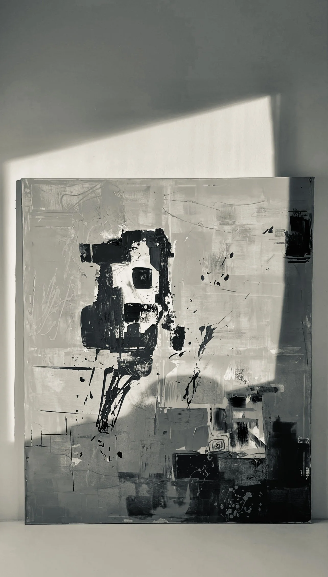 Abstract black, white, and gray painting with various shapes and splatters on a canvas, placed on a white surface with shadows cast on the wall behind.
