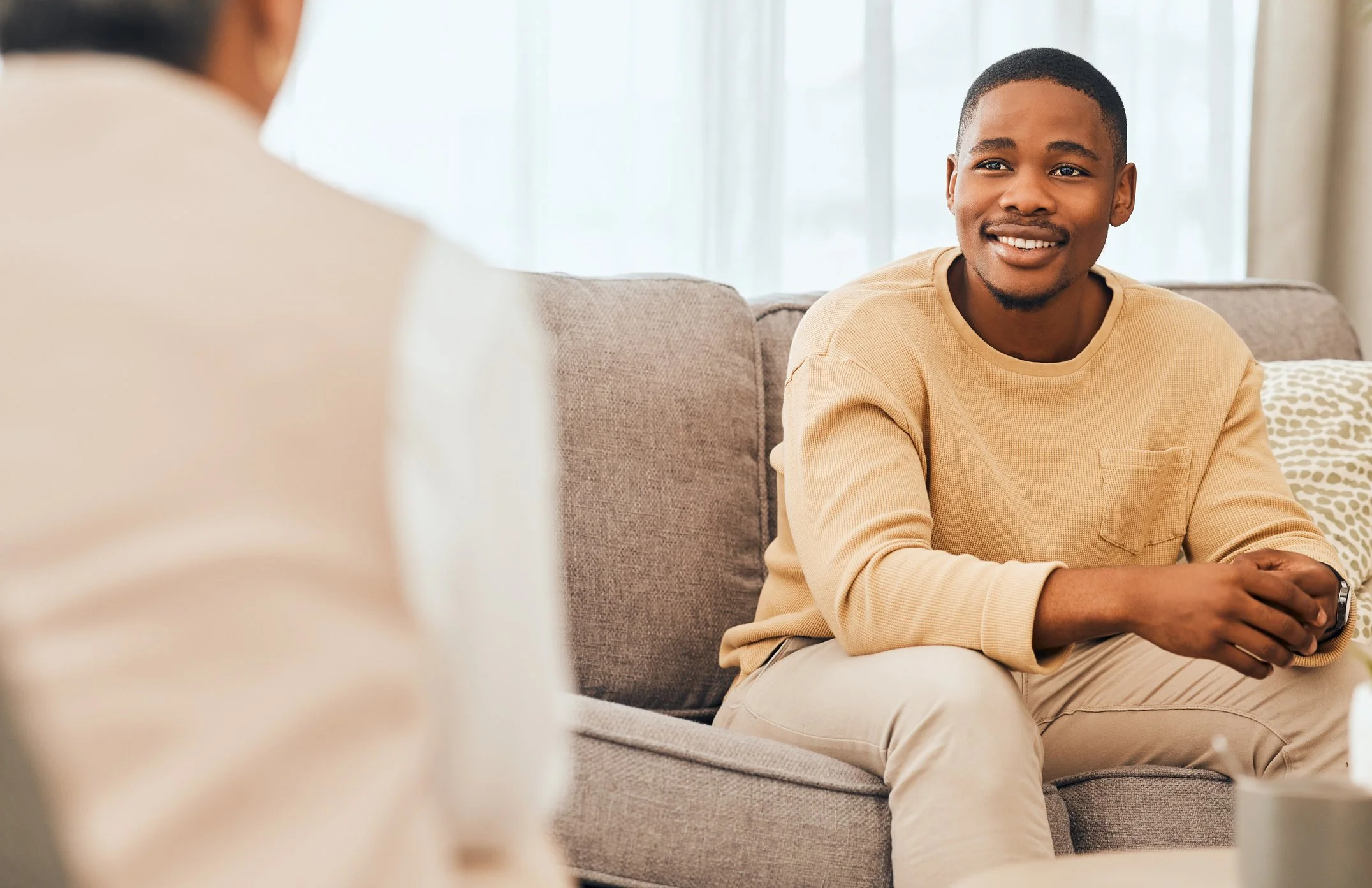 Young man smiling and having a conversation with another person in a cozy, well-lit living room.