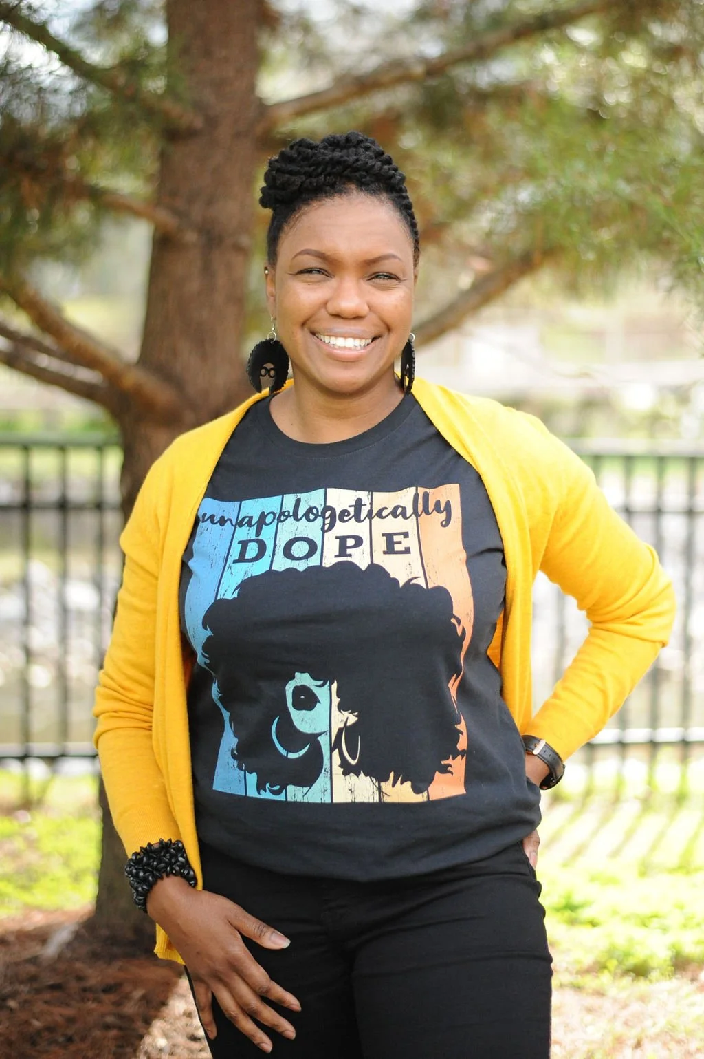 A woman smiling outdoors in front of a tree and fence, wearing a black t-shirt with a graphic of a woman's face with an afro, and a yellow cardigan.