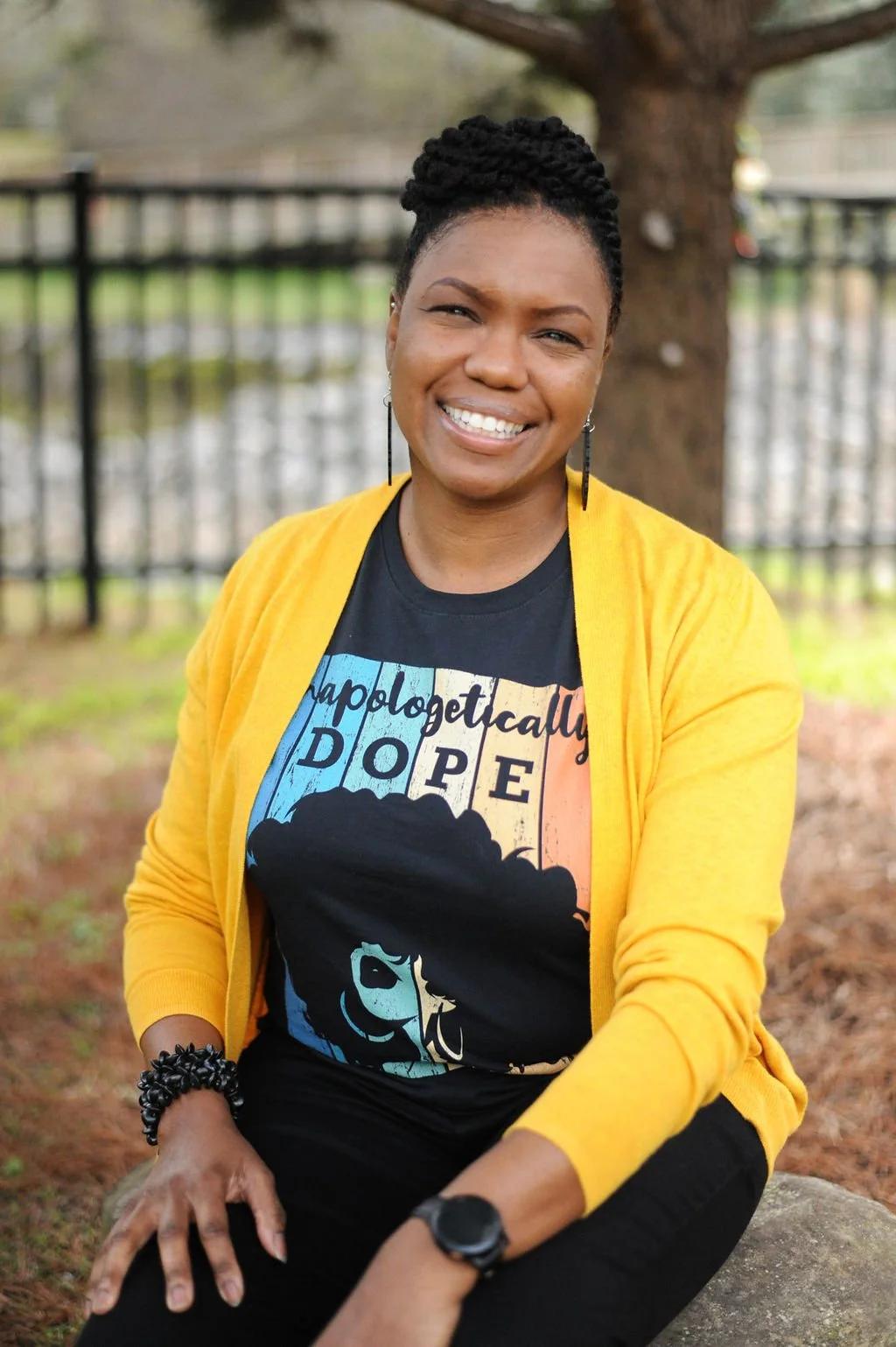 A woman with braided hair smiling while sitting outdoors near a tree and black metal fence, wearing a black graphic t-shirt, yellow cardigan, bracelet, and watch.