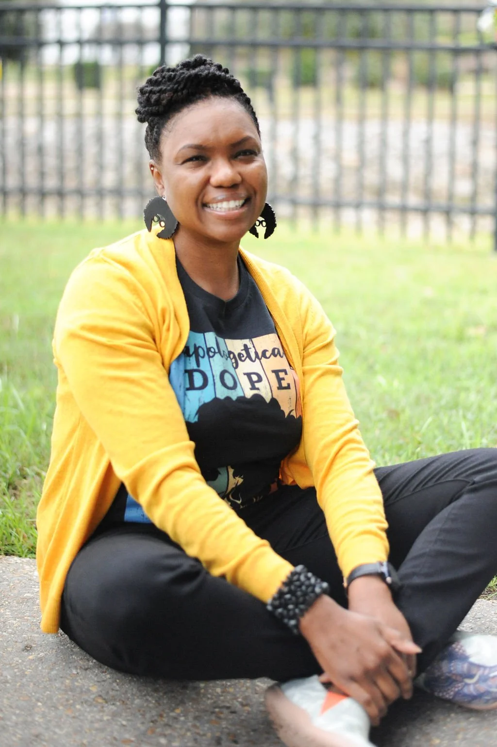 A woman with braided hair, wearing black earrings, a black t-shirt, a yellow cardigan, and black pants, is sitting on the sidewalk outdoors with her legs crossed, smiling at the camera.