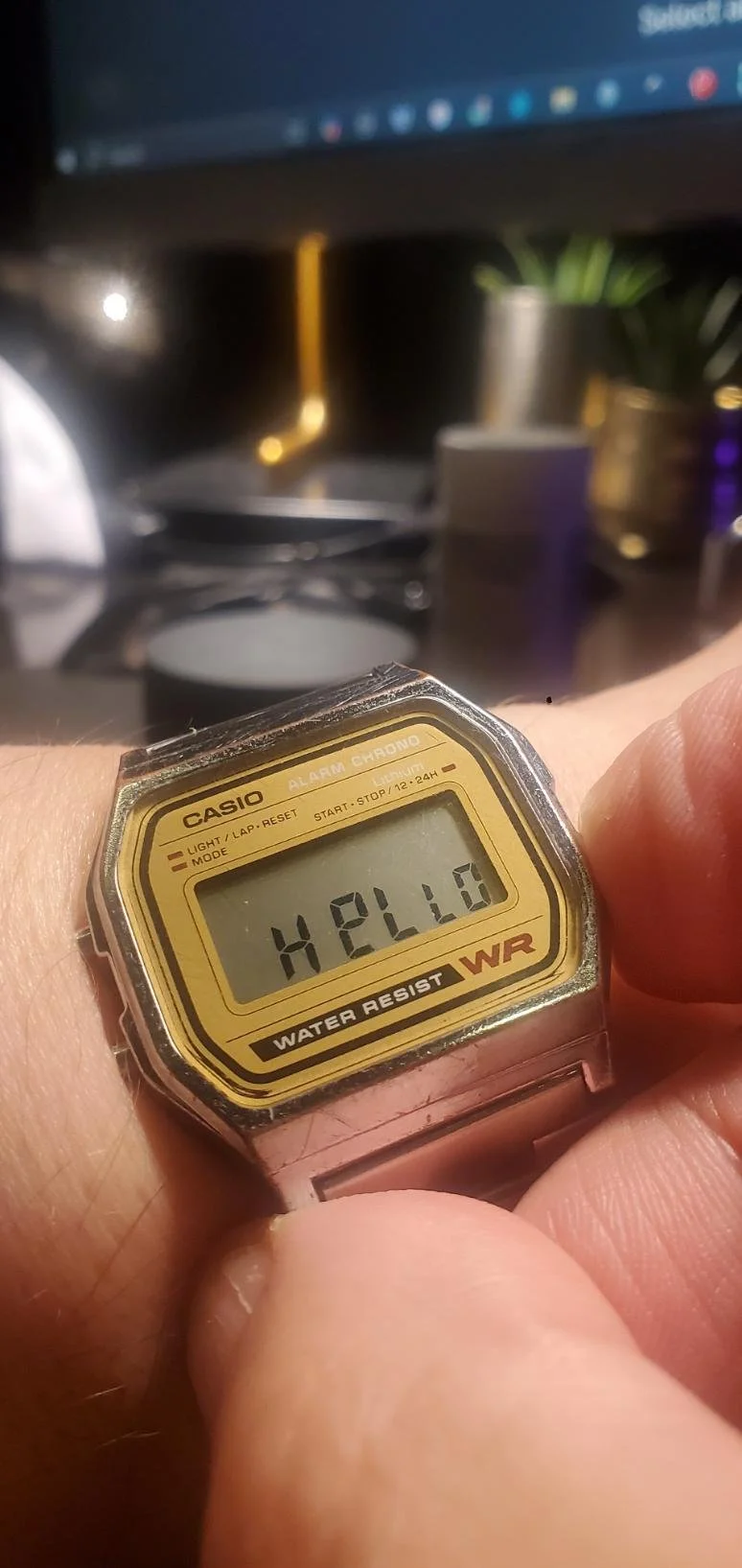 A digital wristwatch displaying the time 4:21:50 with the word 'HELLO' on the screen, held by a person's hand, in front of a blurred background of a computer monitor and various objects.