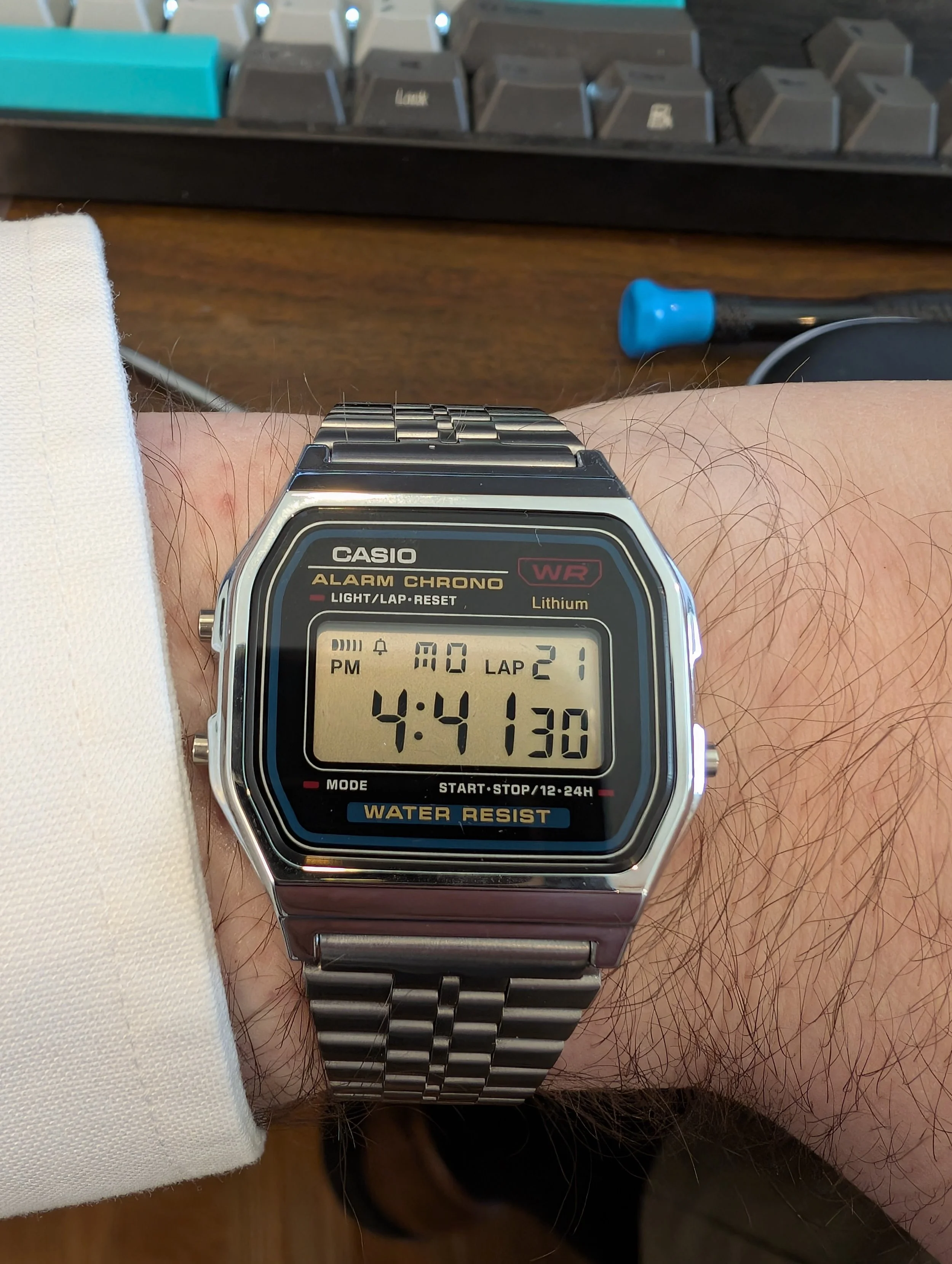 A digital Casio wristwatch with a metal band displaying the time as 4:41:30, on a person's left wrist. The watch face also shows the date as August 21 and the day as Monday, with a lap count of 2. The background includes a wooden desk, partially visible keyboard, and a blue-handled screwdriver.