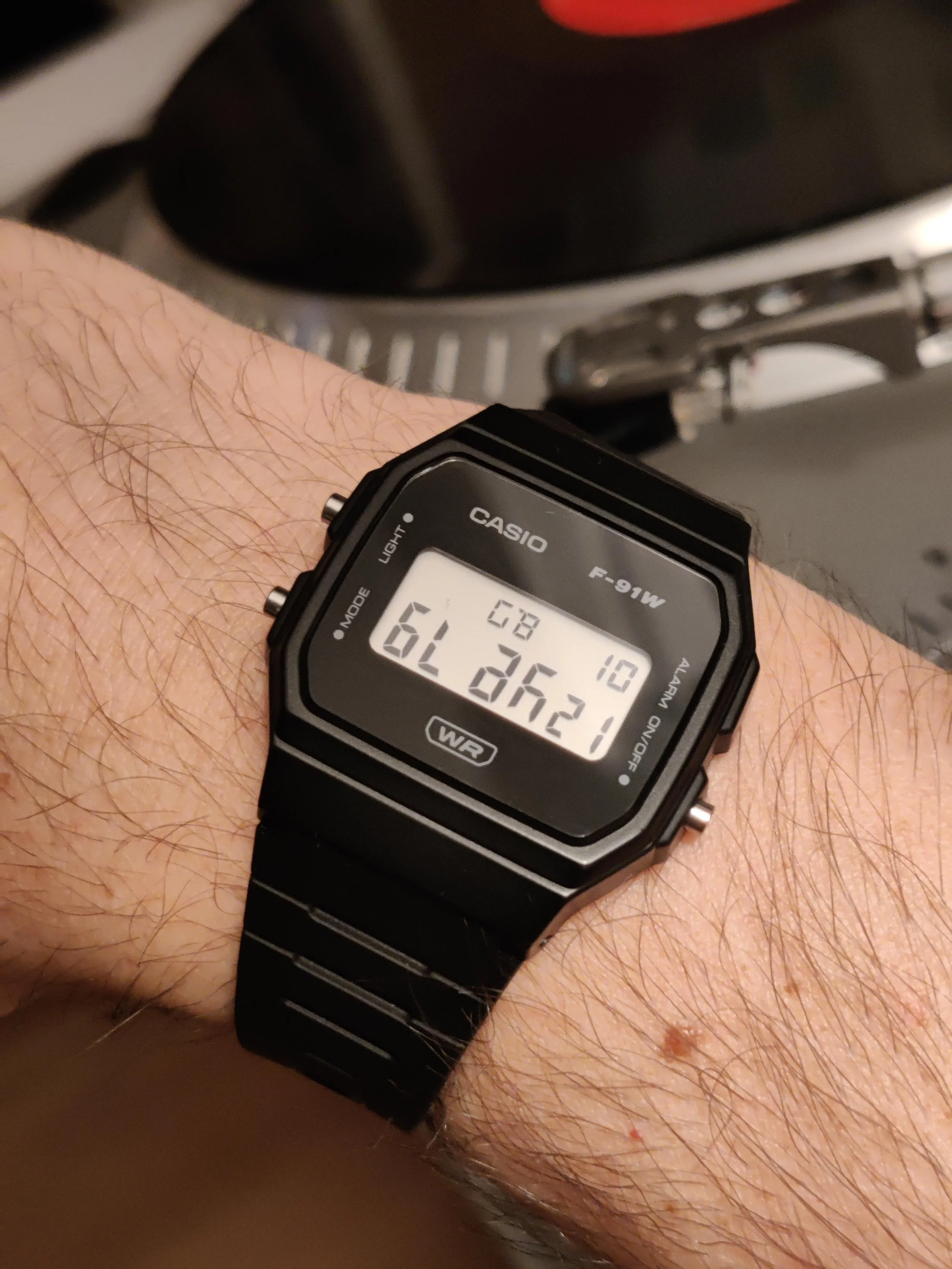 A person's wrist wearing a black Casio digital watch displaying the date and time, with a grill in the background.