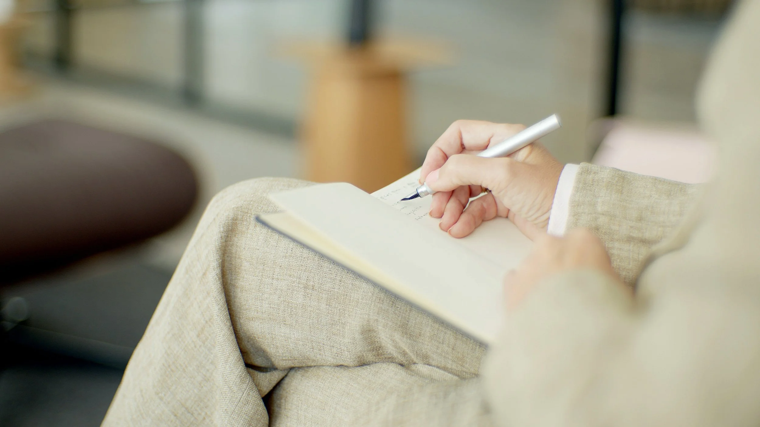 Person writing notes in a notebook while sitting, wearing beige pants and a matching blazer.