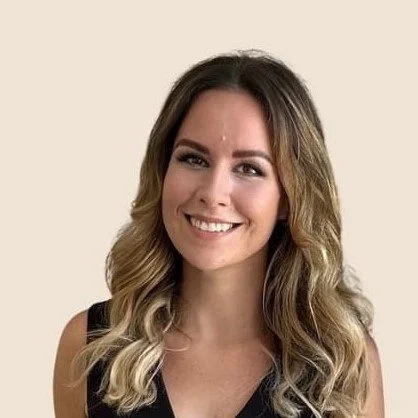 A woman with long wavy brown hair, smiling, wearing a black sleeveless top against a beige background.
