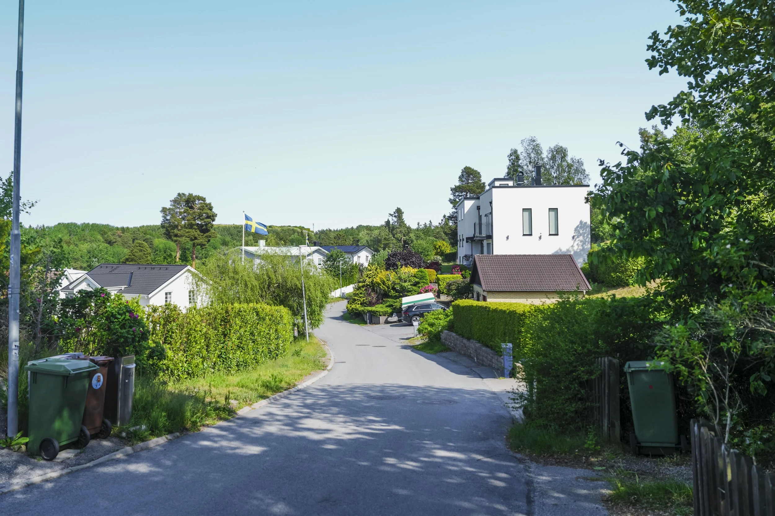 En lugn gata i en svensk förort med gröna träd och buskar, bostadshus och en svensk flagga till vänster, under en klar blå himmel.
