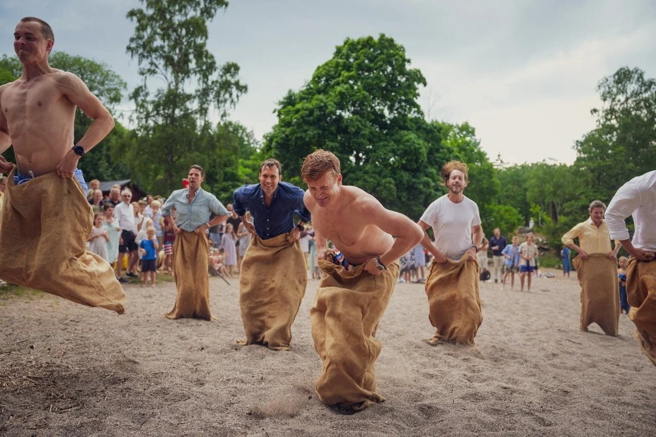 Män tävlar i säckhoppning under en utomhusfestival, omgivna av en publik och gröna träd.