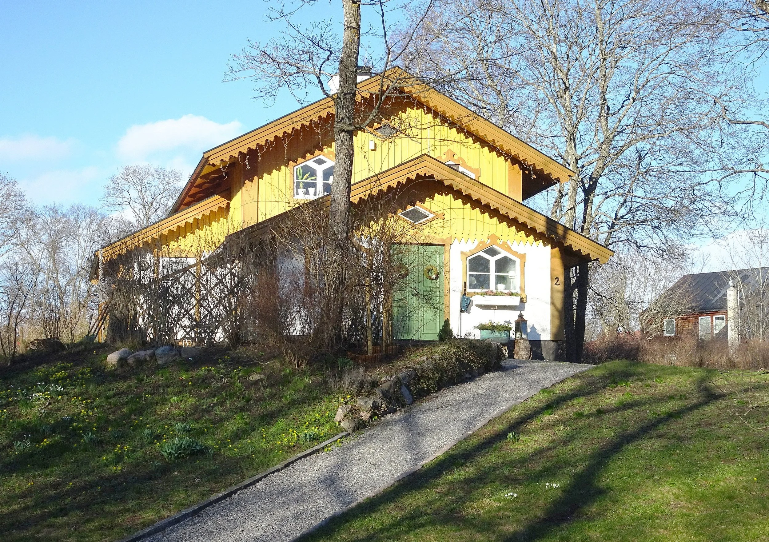 En färgglad hus i trä med gula väggar och snedtak, omgivet av träd och gräs, på en solig dag.
