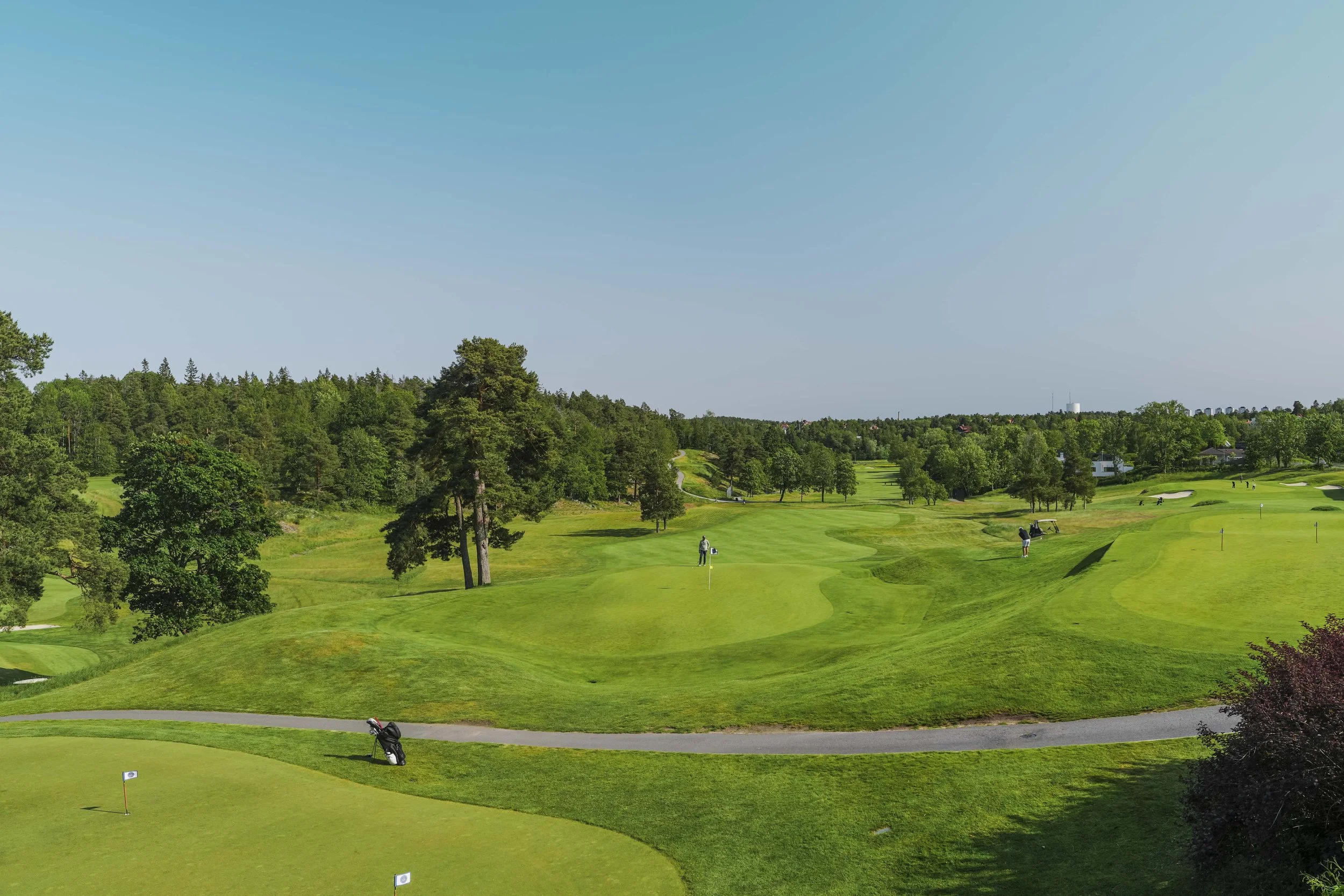 Golfbana med gröna fairways, golfhockeyer och träd i bakgrunden under en klar, blå himmel.