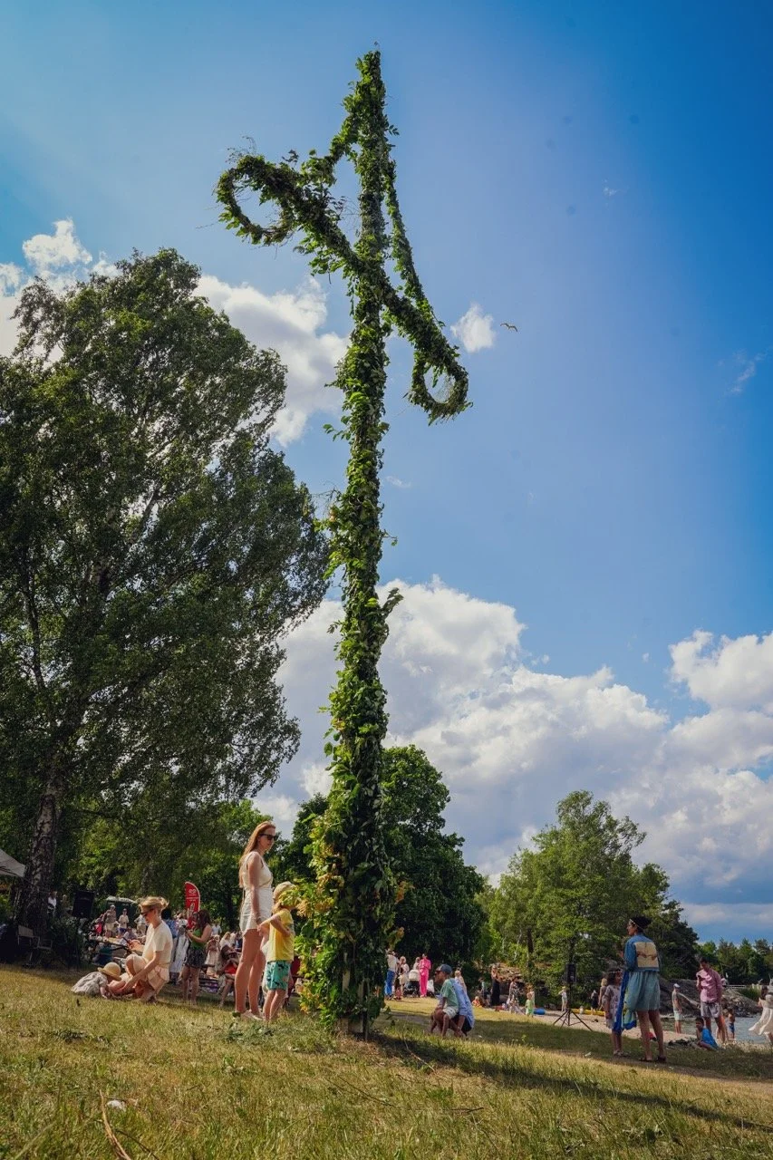 En grodfylt trästam står uppretad i ett parkområde under en klarblå himmel med några moln, omgiven av människor som deltagit i en utomhusaktivitet.