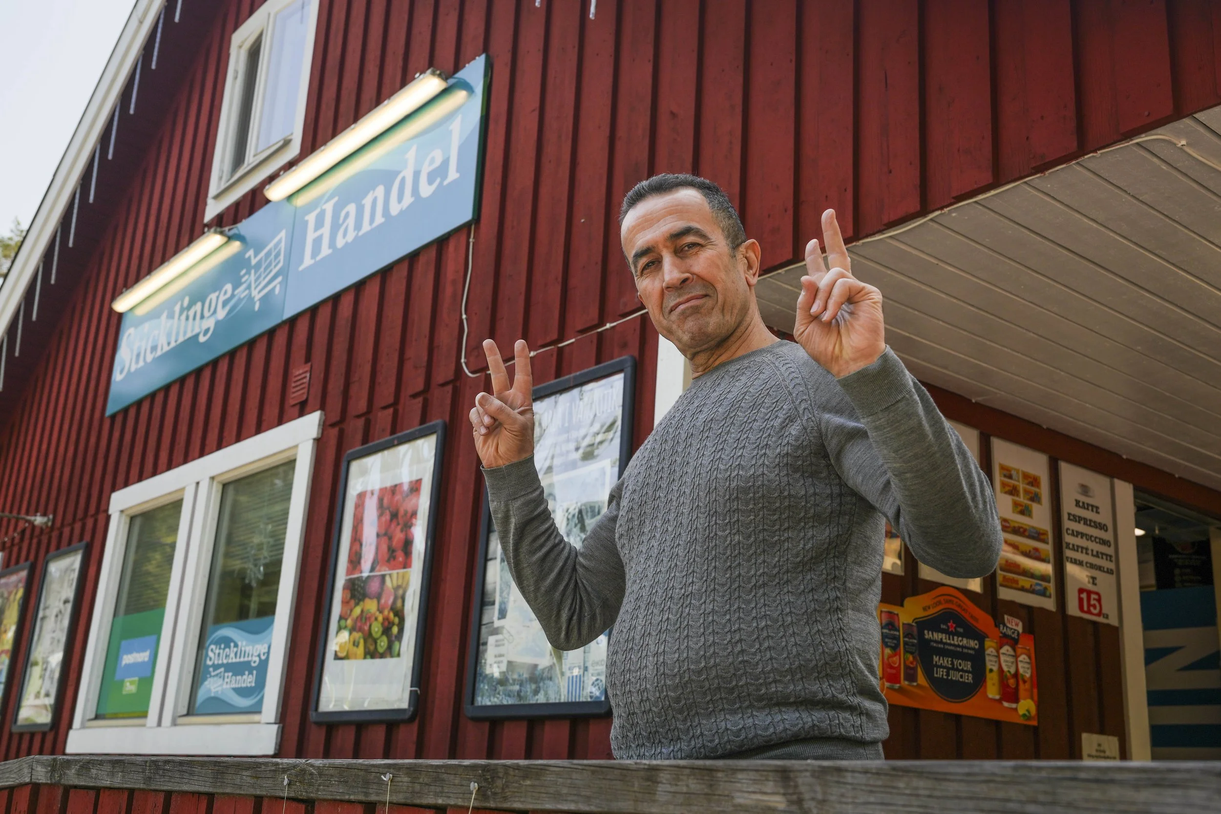 Man i grå tröja gör peace-tecken framför en röd byggnad med klädsamlande affisch och skylt som säger Sticklinge Handel.