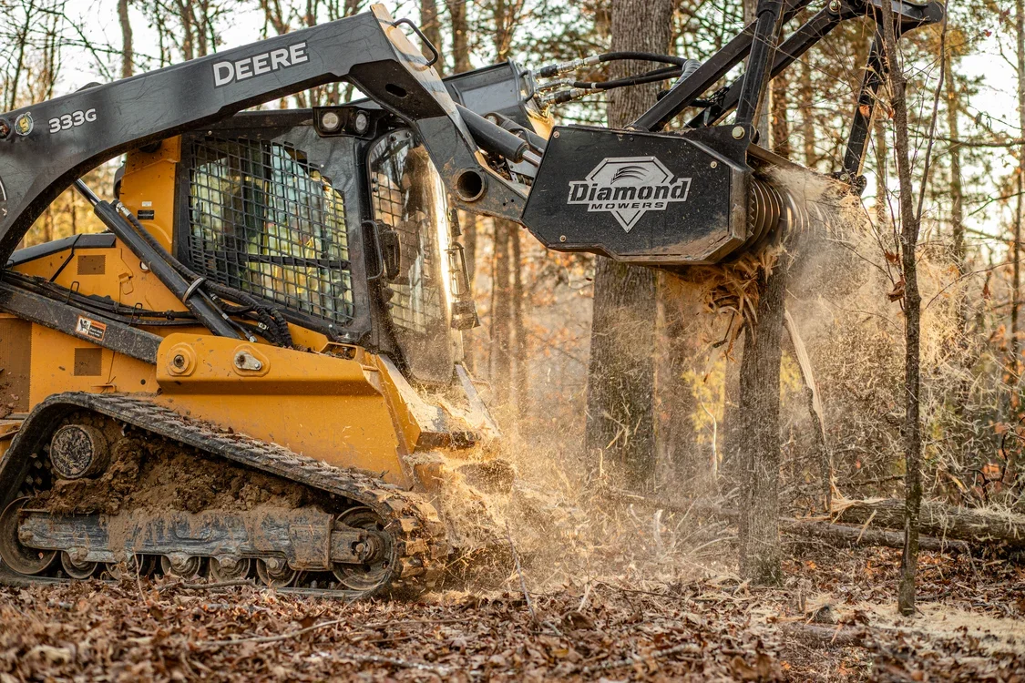A yellow skid steer loader clearing fallen leaves and debris from a wooded area using a hydraulic attachment.