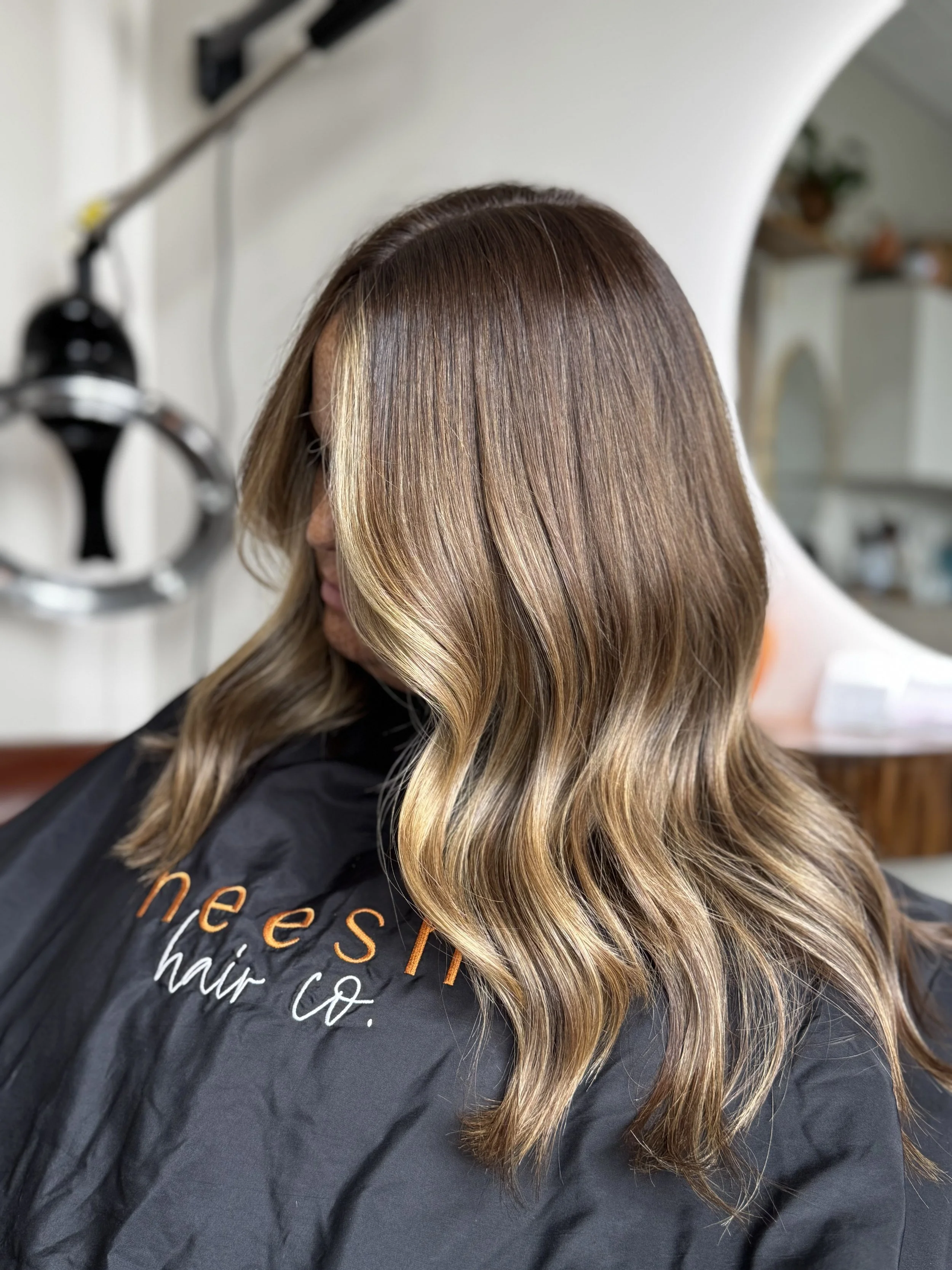 Photo of a woman with wavy, shoulder-length hair with balayage highlights, sitting in a hair salon with a black cape that has orange and white text.
