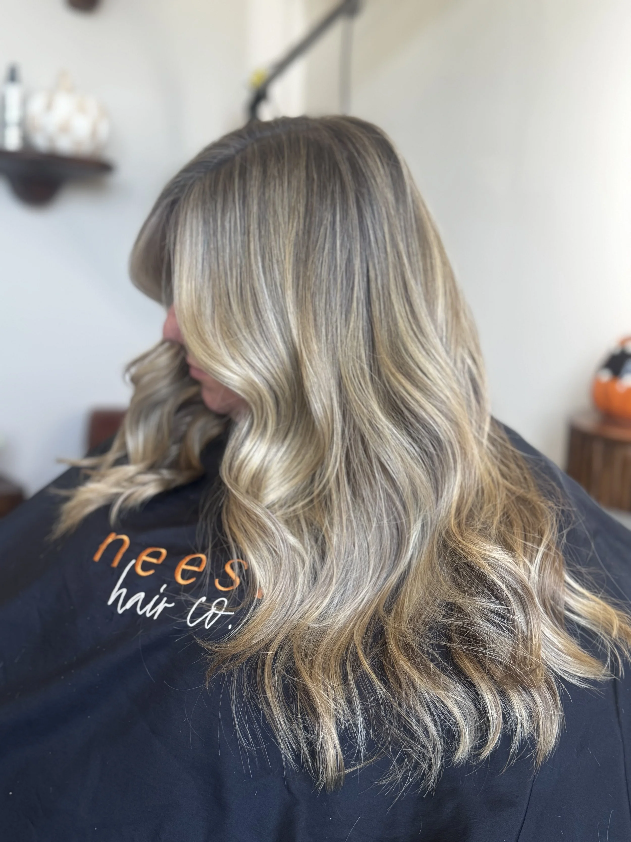 Woman with wavy blonde hair in a salon cape with orange lettering, indoors with blurred shelves and decorations in the background.