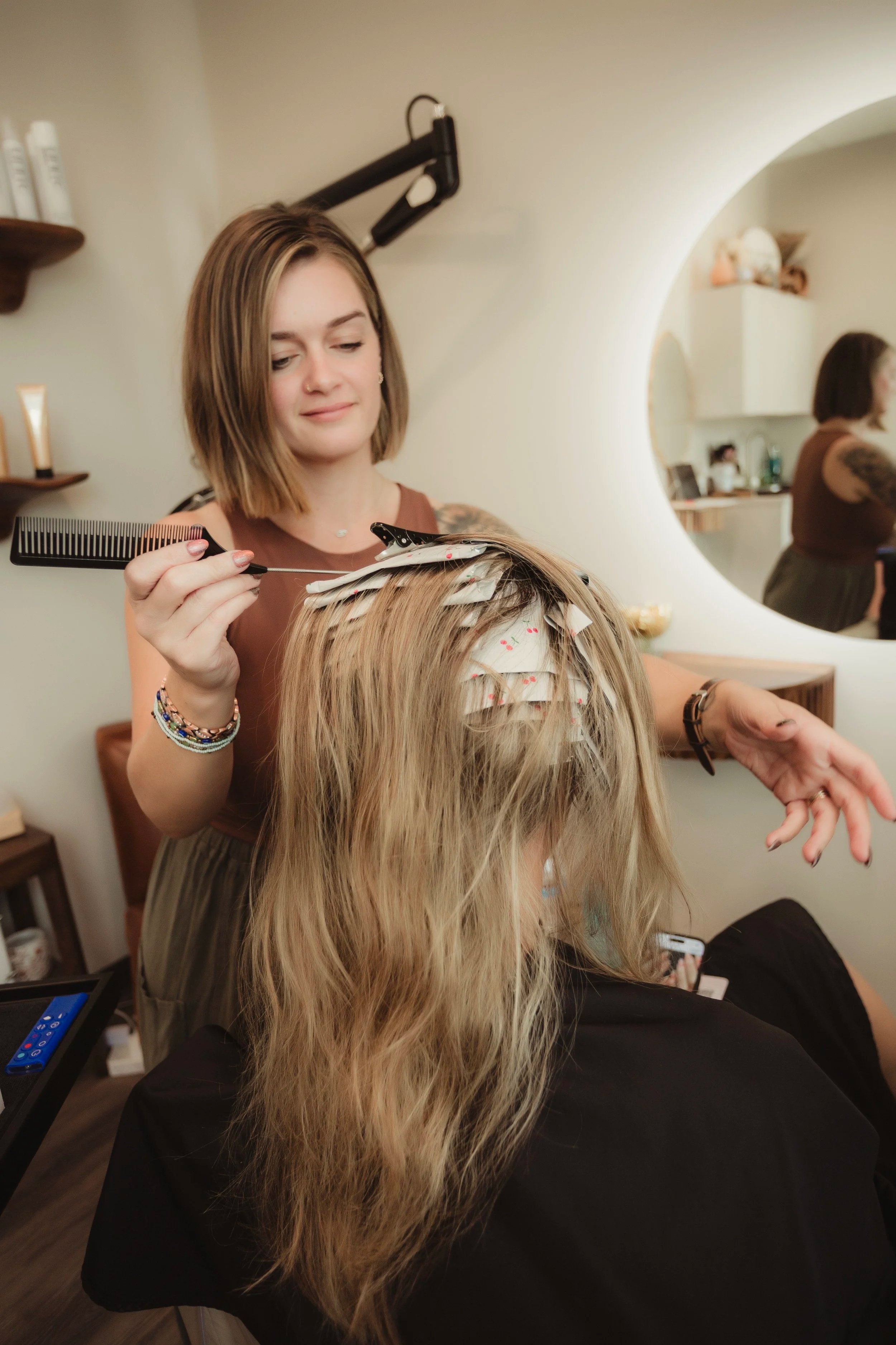 A hairdresser with shoulder-length brown hair and a sleeveless top clips hair extensions into a woman's long blonde hair in a salon.