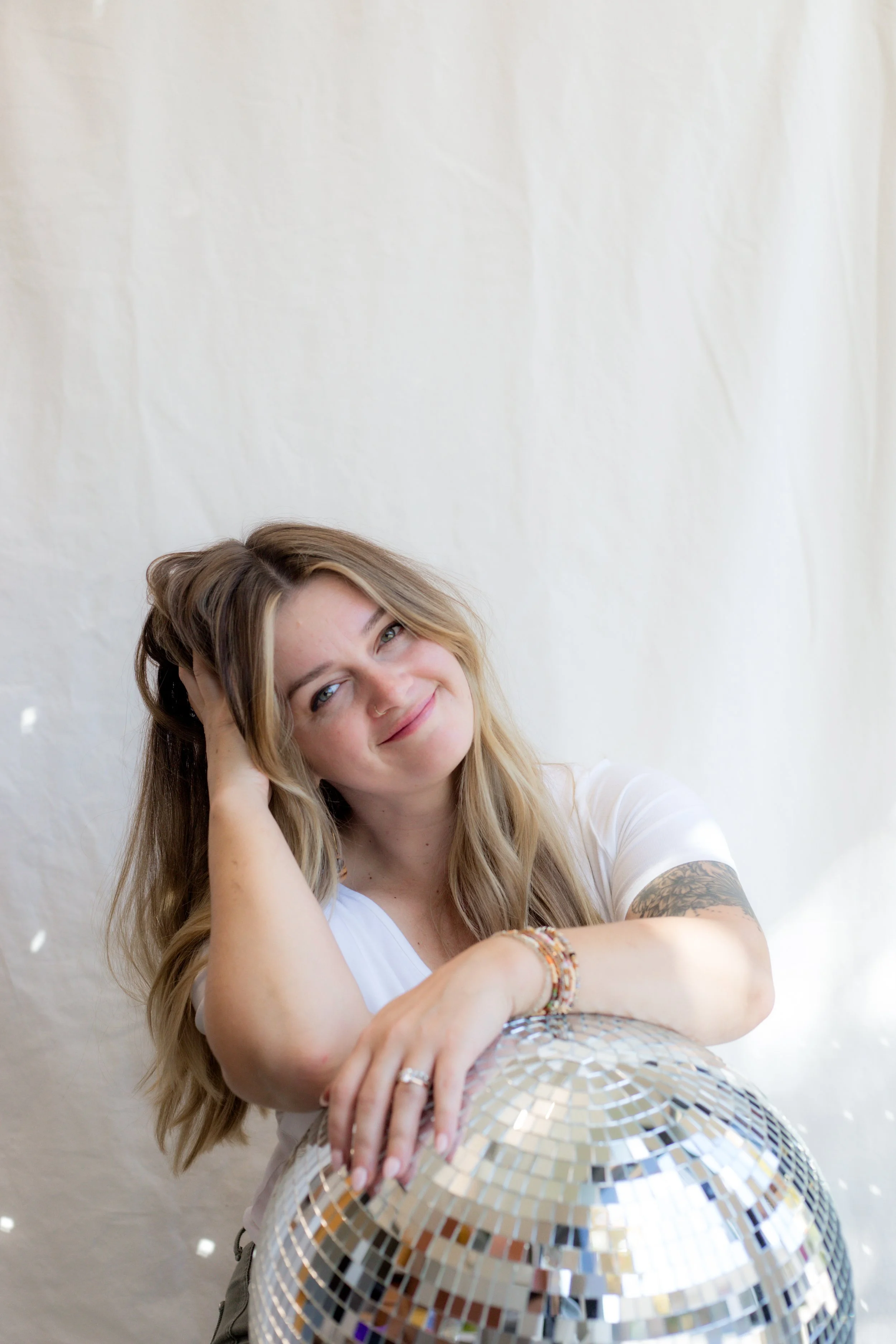 A woman with long blonde hair smiling, resting her head on her hand, leaning on a large disco ball.
