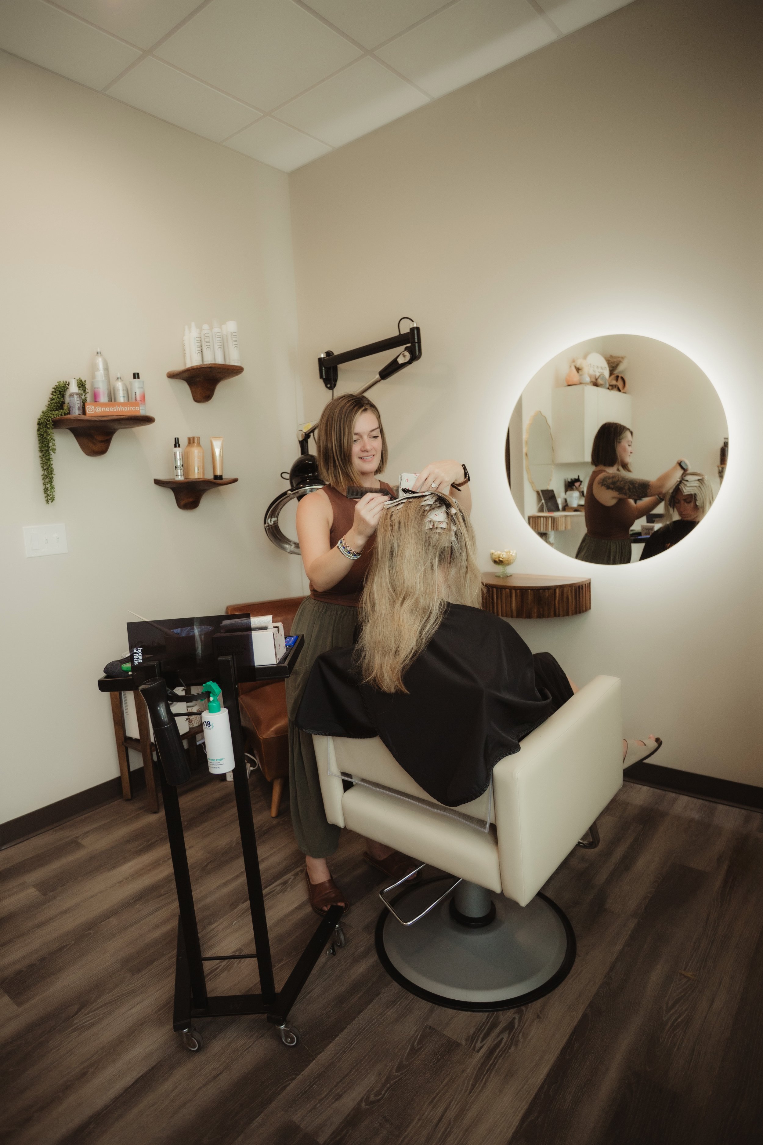 A woman getting her hair styled at a salon, sitting in a white salon chair, with a stylist standing behind her, using hair coloring tools, reflected in a round mirror on the wall.