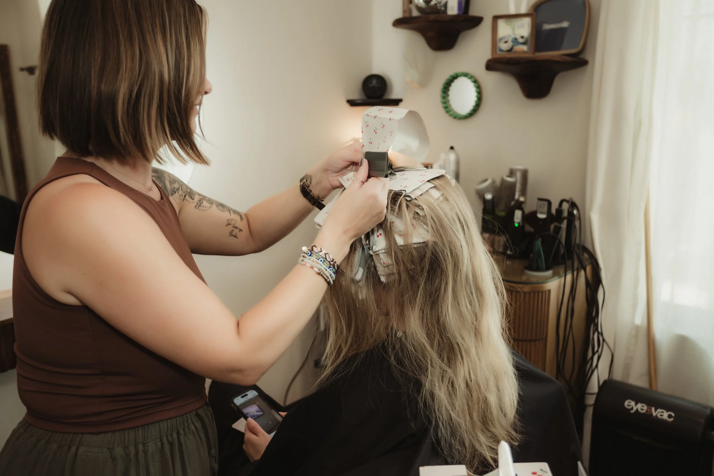 A woman is applying hair color or highlights to a client's long, blonde hair using foils and a foil applicator in a hair salon. The client is sitting in a salon chair with her head slightly tilted forward.