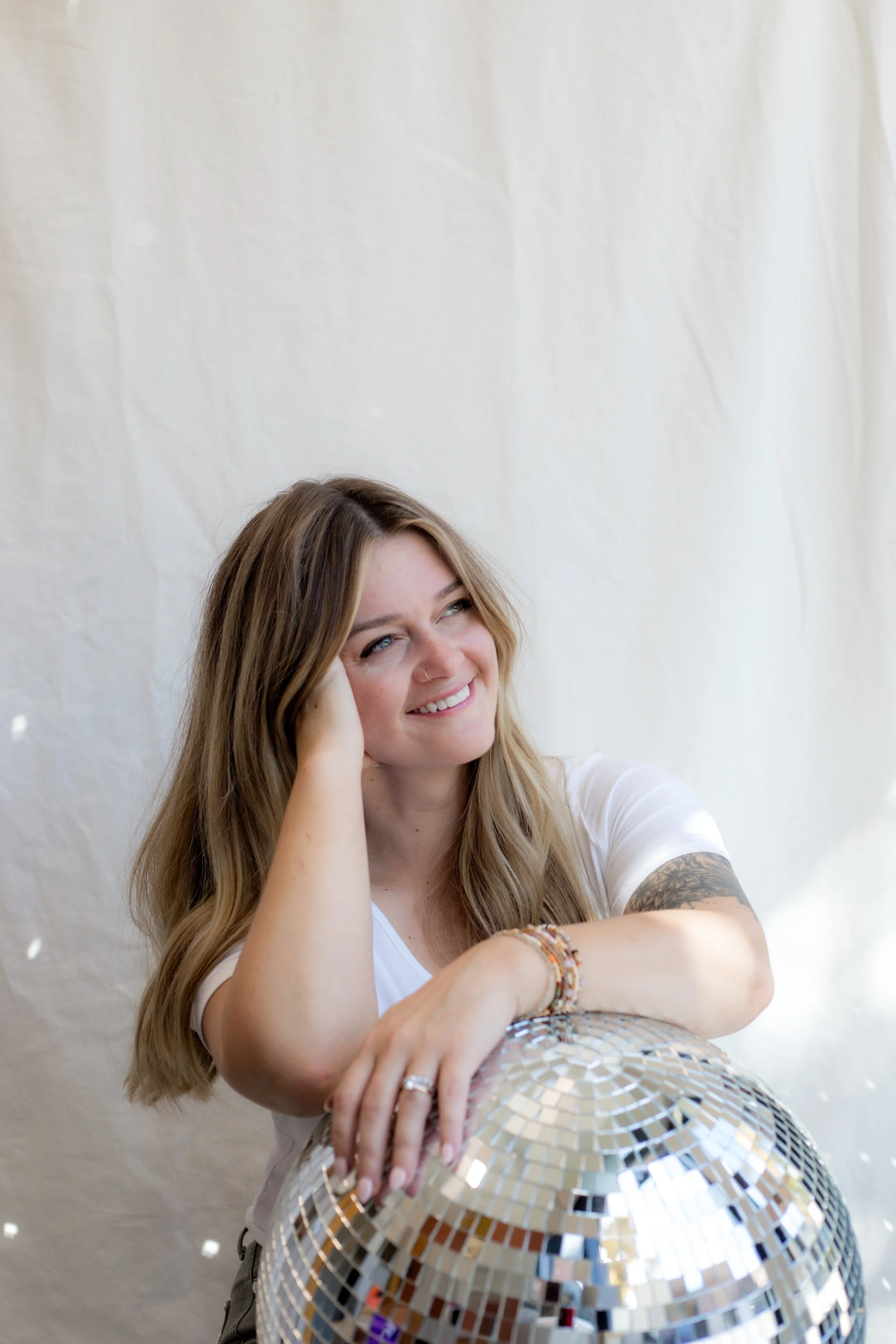 A woman with long wavy hair, a nose ring, wearing a white t-shirt, and multiple bracelets, leaning on a large disco ball with a white background.