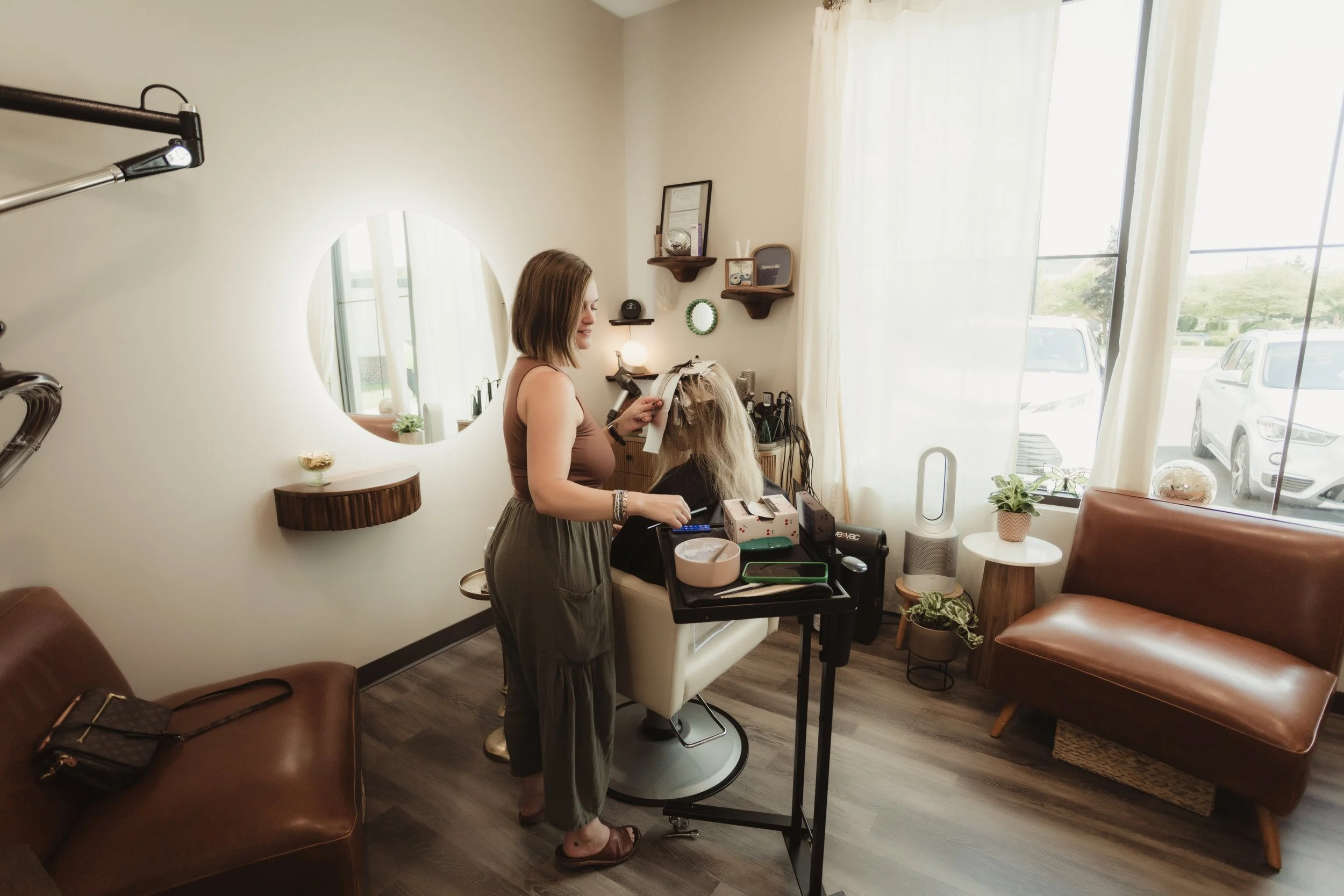 A woman getting her hair styled at a salon, with hairdresser using tools at a styling station near a large window with sheer curtains. Decor includes a mirror, small plants, and shelves on the wall.