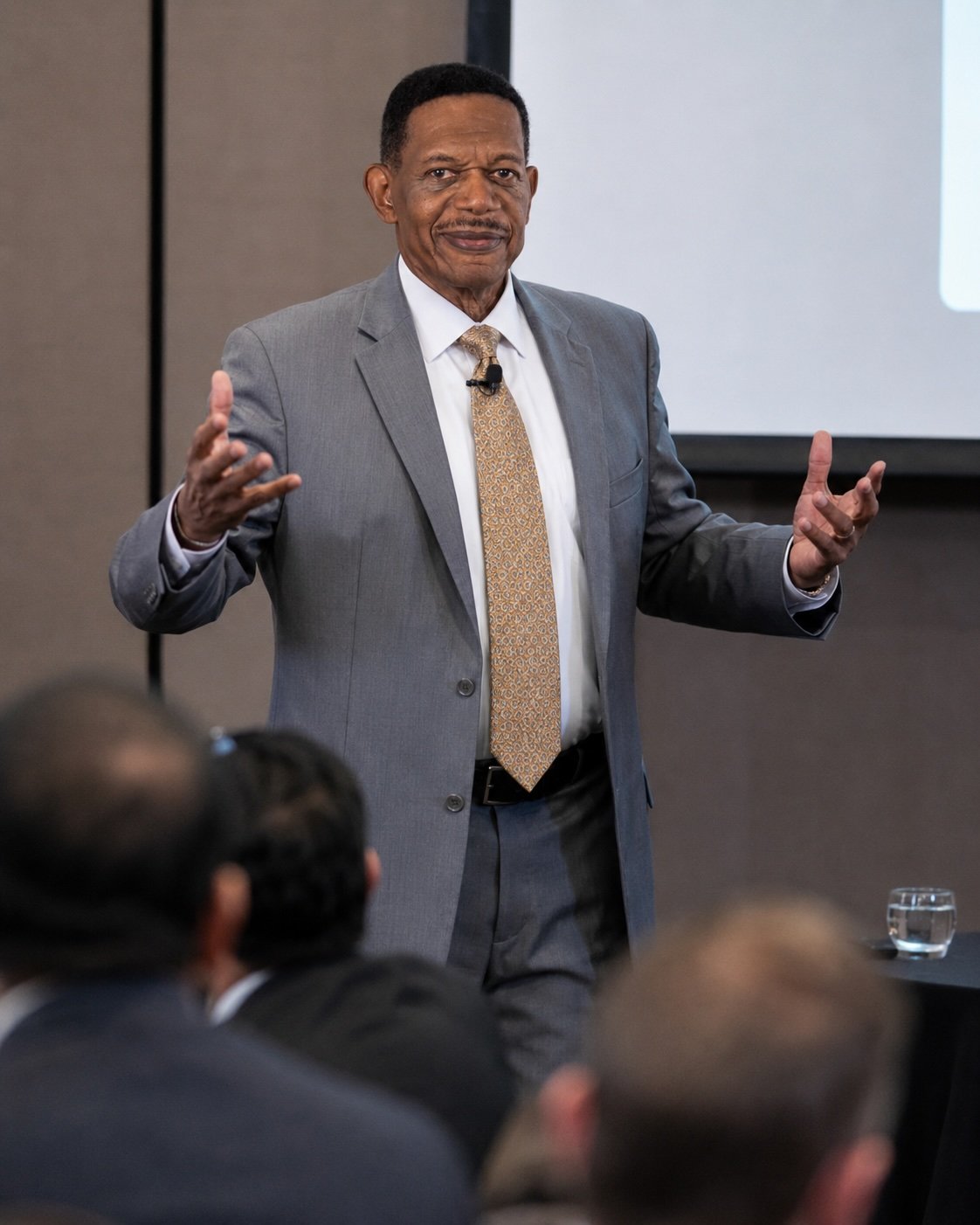Brandon Campbell, in a gray suit, white shirt, and patterned tie, is giving a presentation to an audience in a conference room for The Advisory Commission.