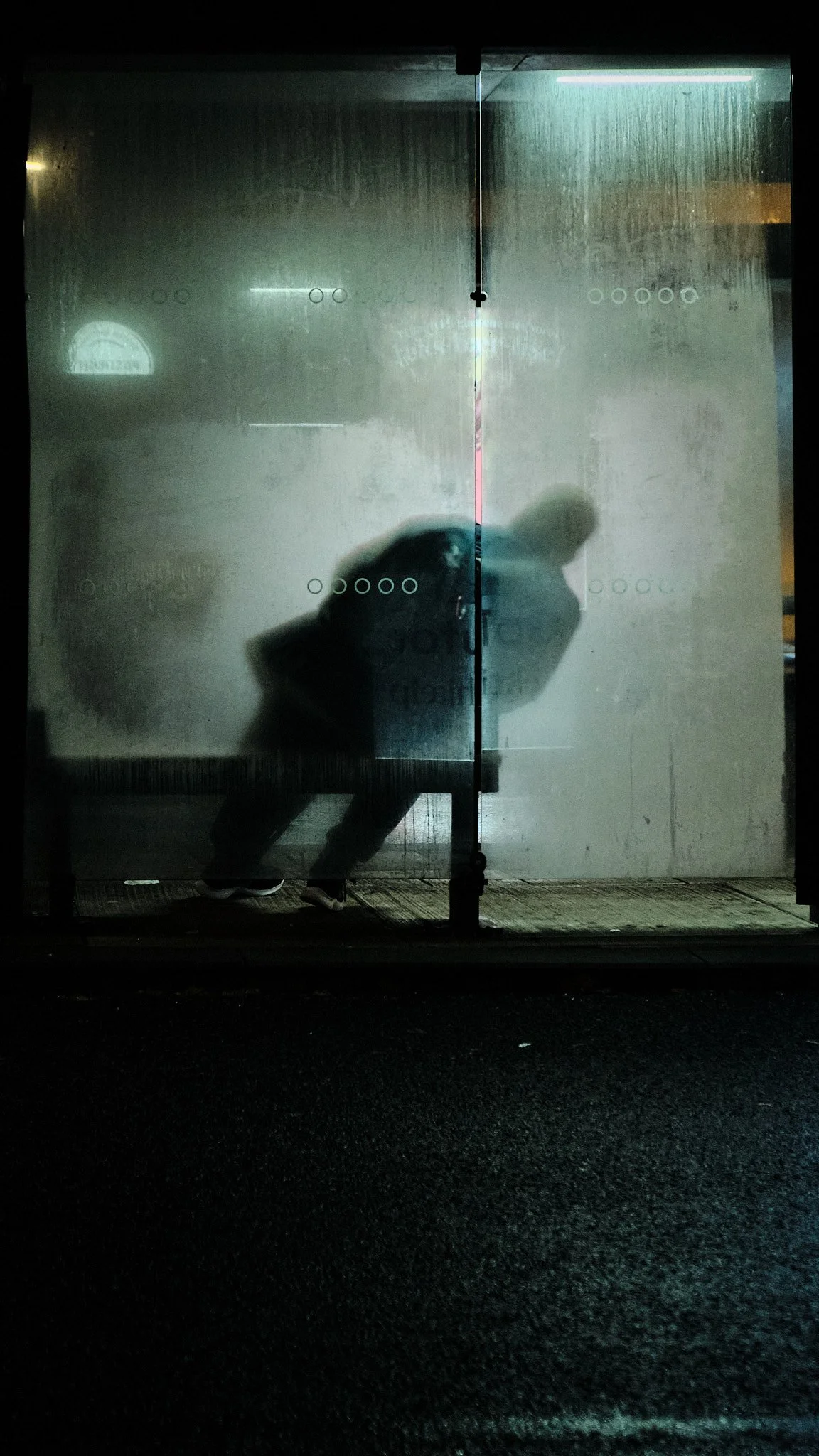Silhouette of a person leaning over inside a foggy glass bus stop shelter at night.