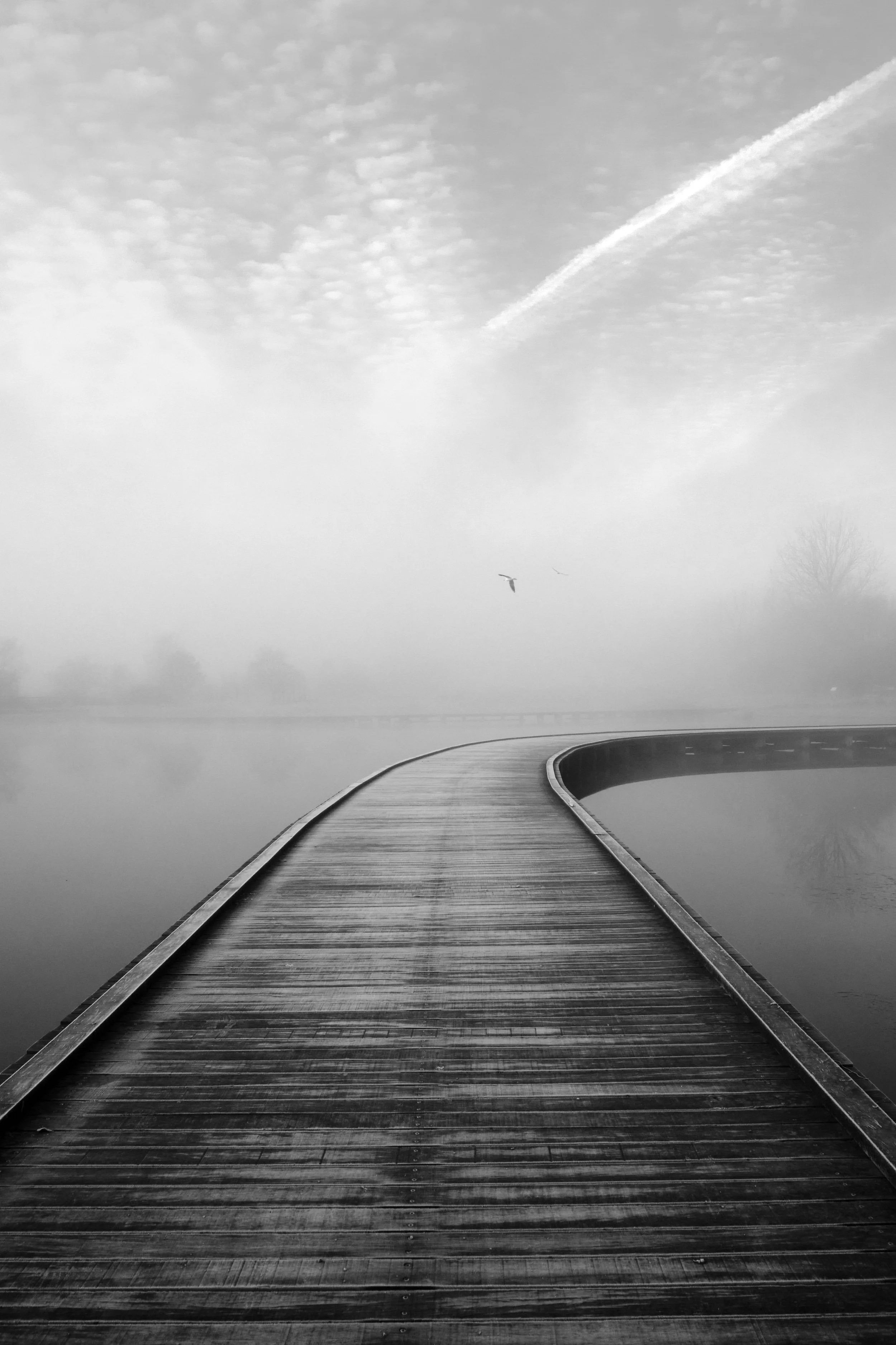 A black and white photo of a curved wooden bridge over a foggy body of water with a bird flying in the distance and clouds in the sky.