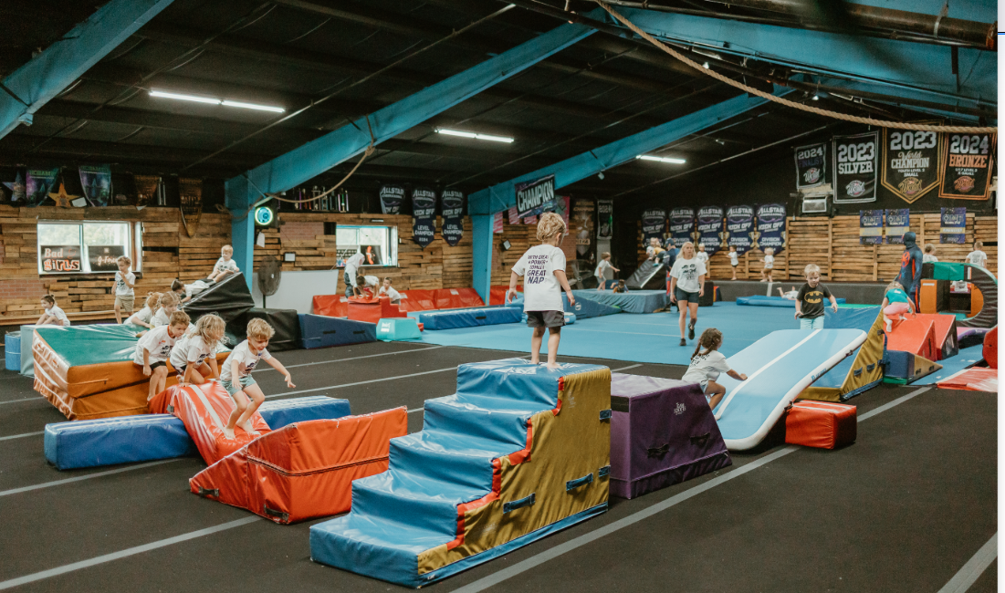 Children playing on inflatable bounce houses and slides inside an indoor play area with banners and trophies hanging on the walls.