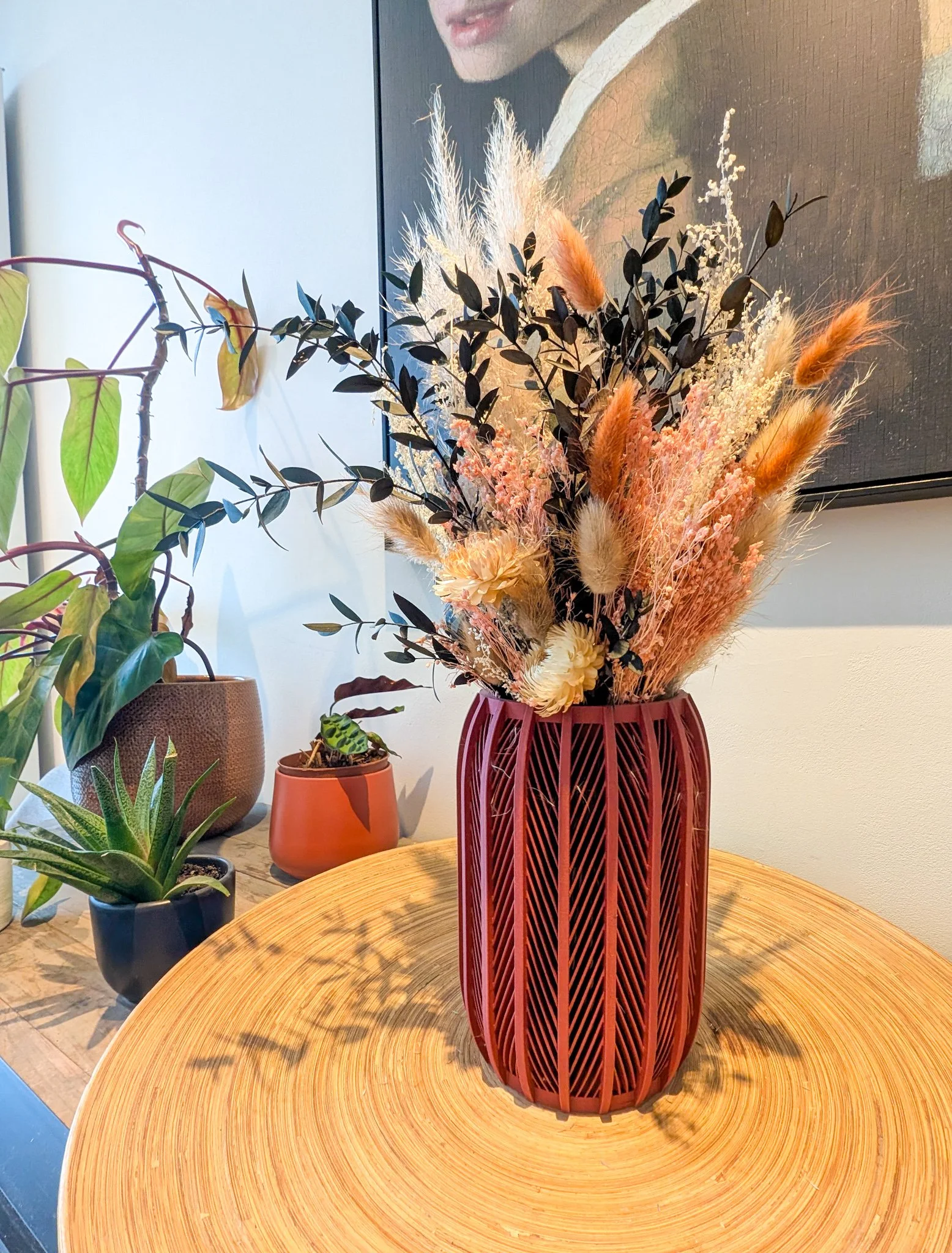 Decoratieve vaas met gedroogde bloemen en planten op een ronde houten tafel, omringd door andere kamerplanten.