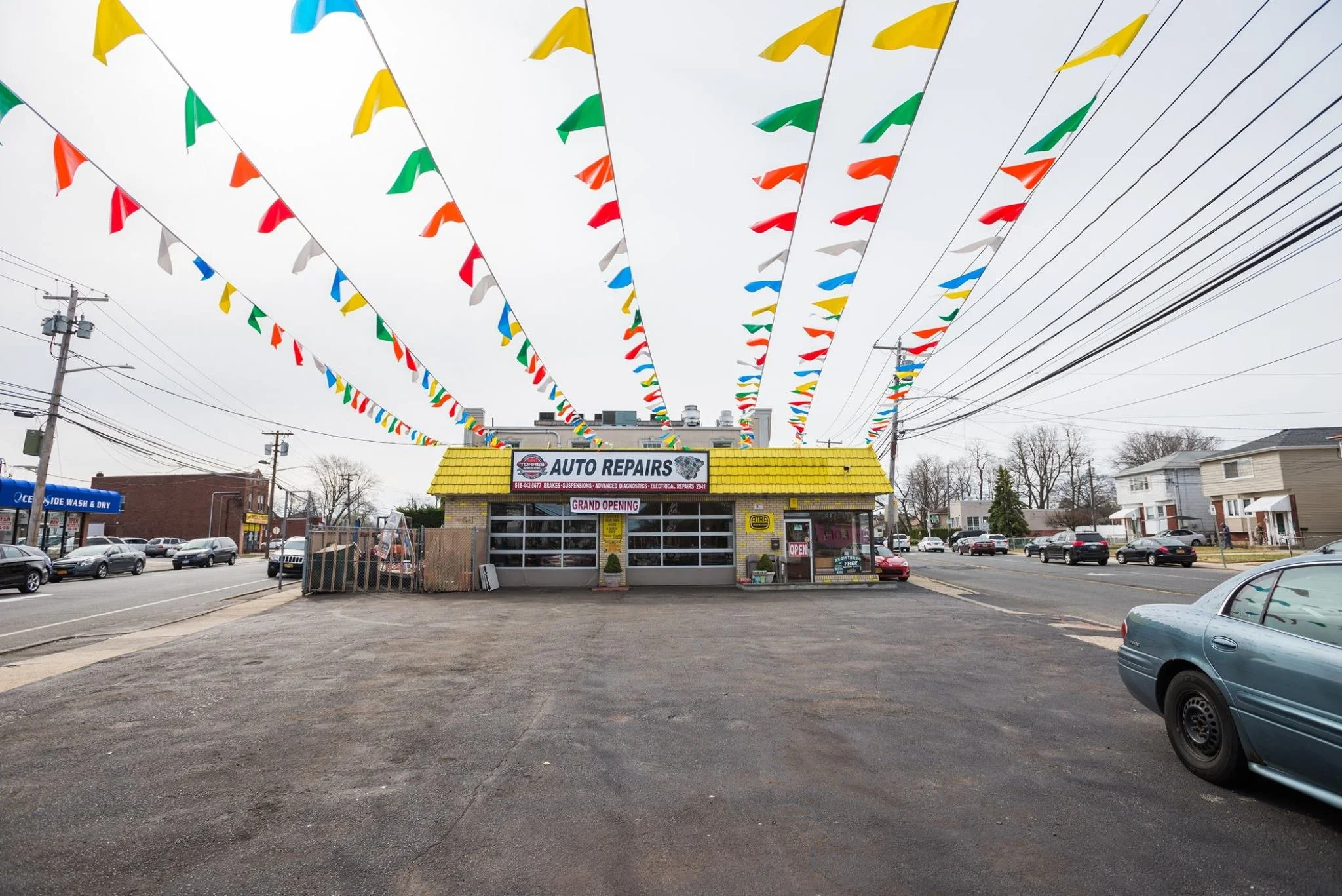 A small auto repair shop with a bright yellow roof and a sign advertising brakes, suspensions, diagnostics, and electrical repairs, located in a busy parking lot with cars and power lines overhead, decorated with colorful pennant flags hanging overhead.