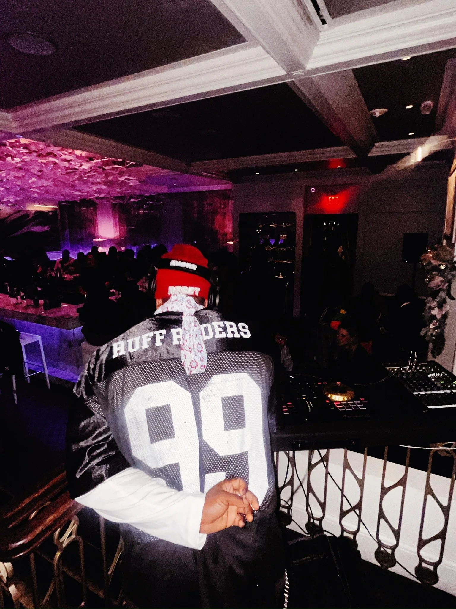DJ performing at a nightclub with a crowd in the background. The DJ is wearing a jersey with the word 'RUFF RIDERS' and the number 99, a red hat, and a bandana. The nightclub has dark lighting with pink and purple lights.