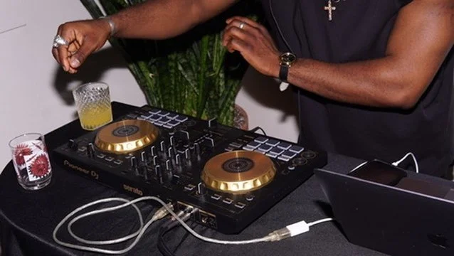 Close-up of a DJ mixing music with a Pioneer DJ controller on a black table, showing two hands adjusting controls, with drinks and a laptop nearby.