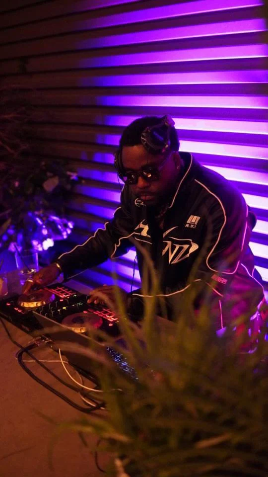 A DJ wearing sunglasses and a jacket, operating a DJ controller with colorful lights, in front of a striped wall illuminated with purple and pink lights.