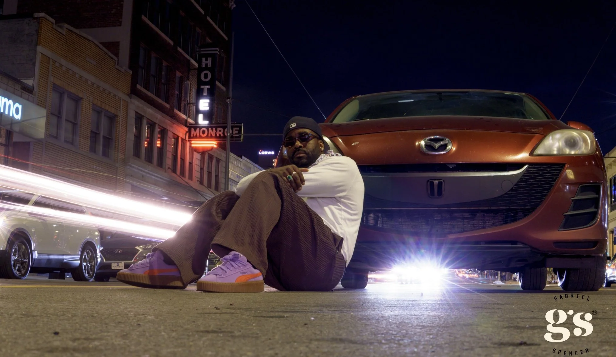 A man with sunglasses, a cap, white shirt, and brown pants sitting on the street at night next to a red Mazda car with illuminated headlights. The background shows city buildings with illuminated signs, including one that says "HOTEL" and another that says "MONROE." Light streaks from passing cars are visible in the scene.
