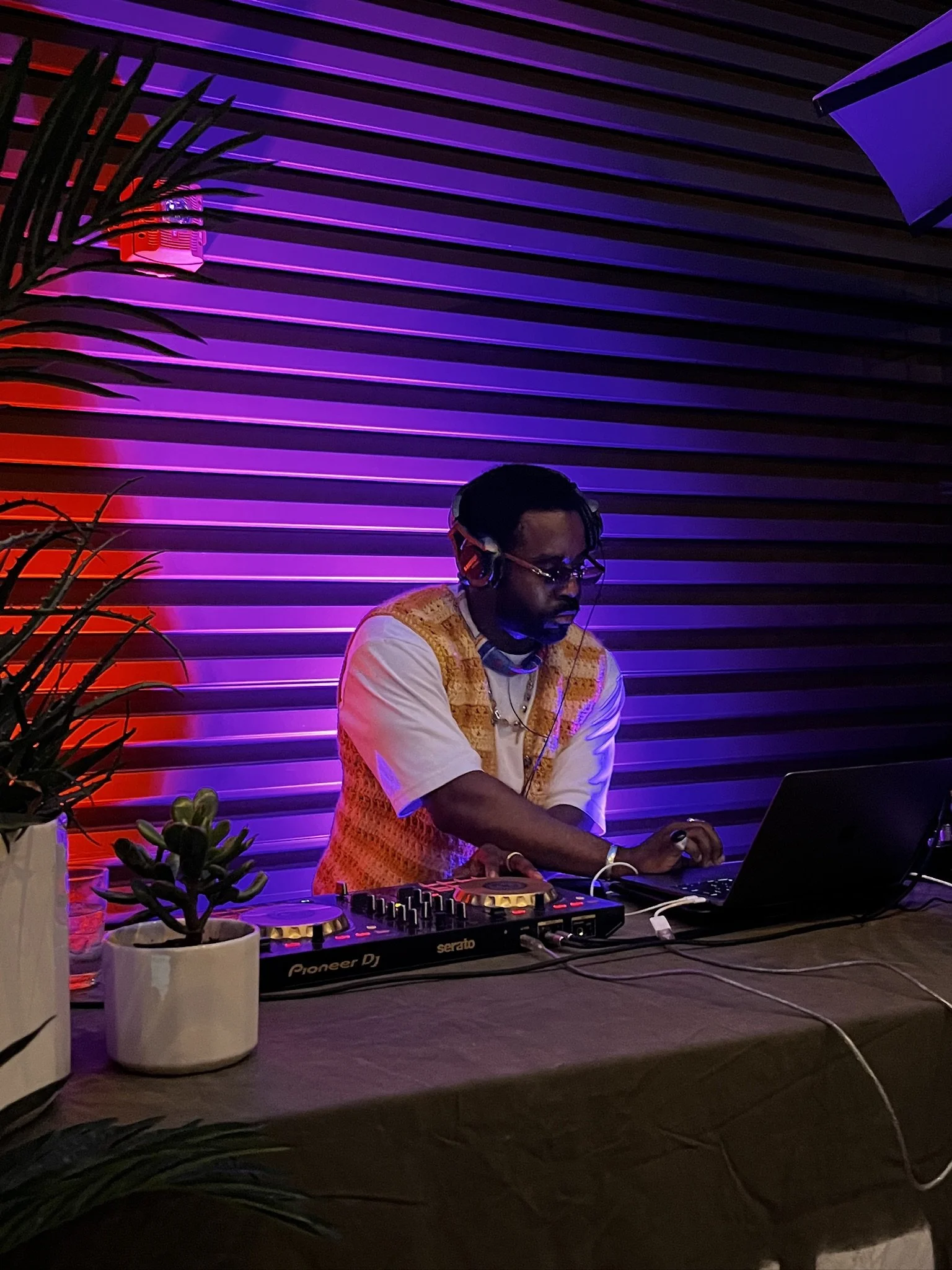 A DJ is working at a setup with a laptop and DJ controller, wearing headphones, in front of a purple lit wall, with potted plants on the table.