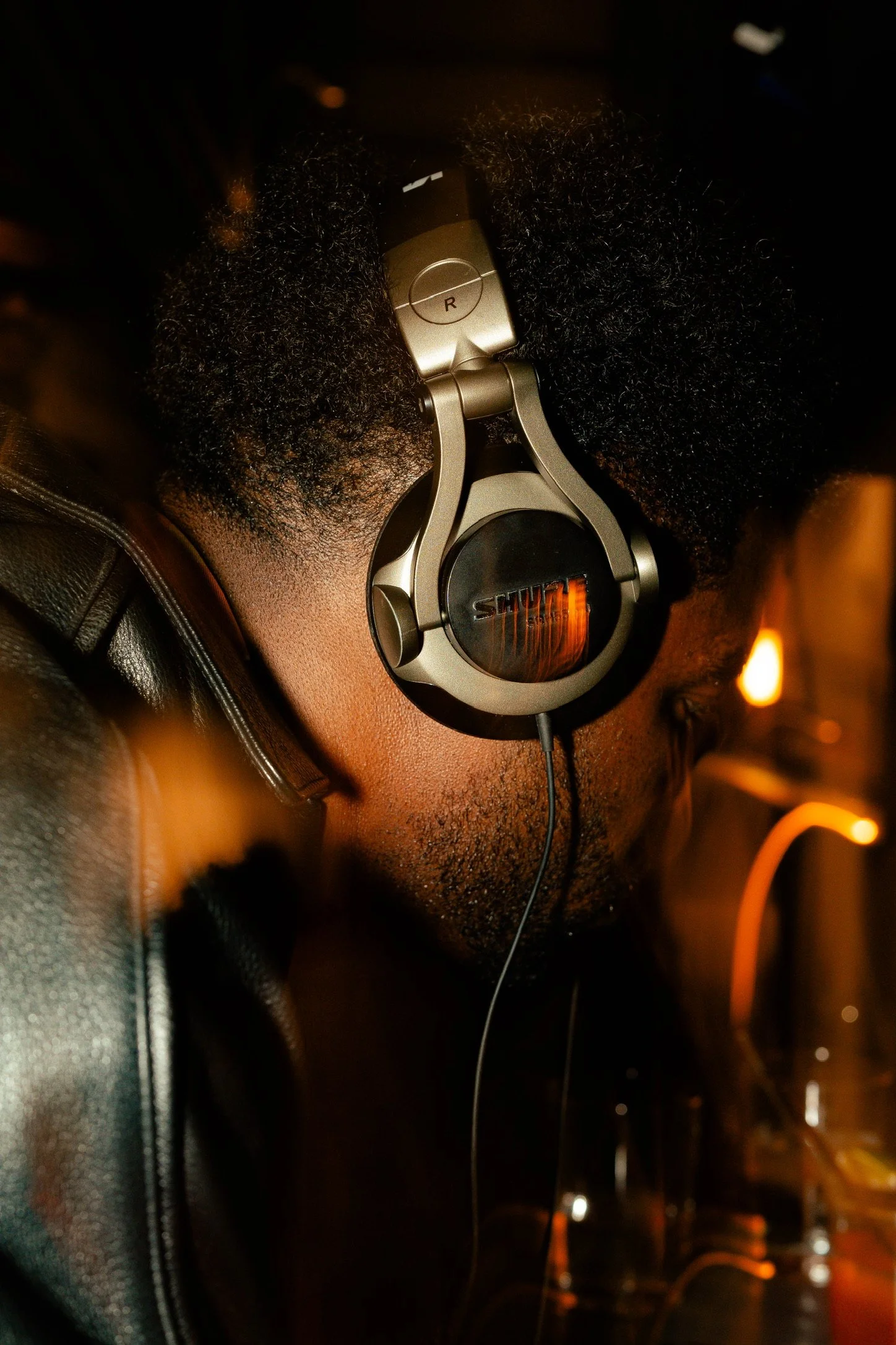 A person with short curly hair wearing large over-ear headphones and a leather jacket, sitting in a dimly lit environment.