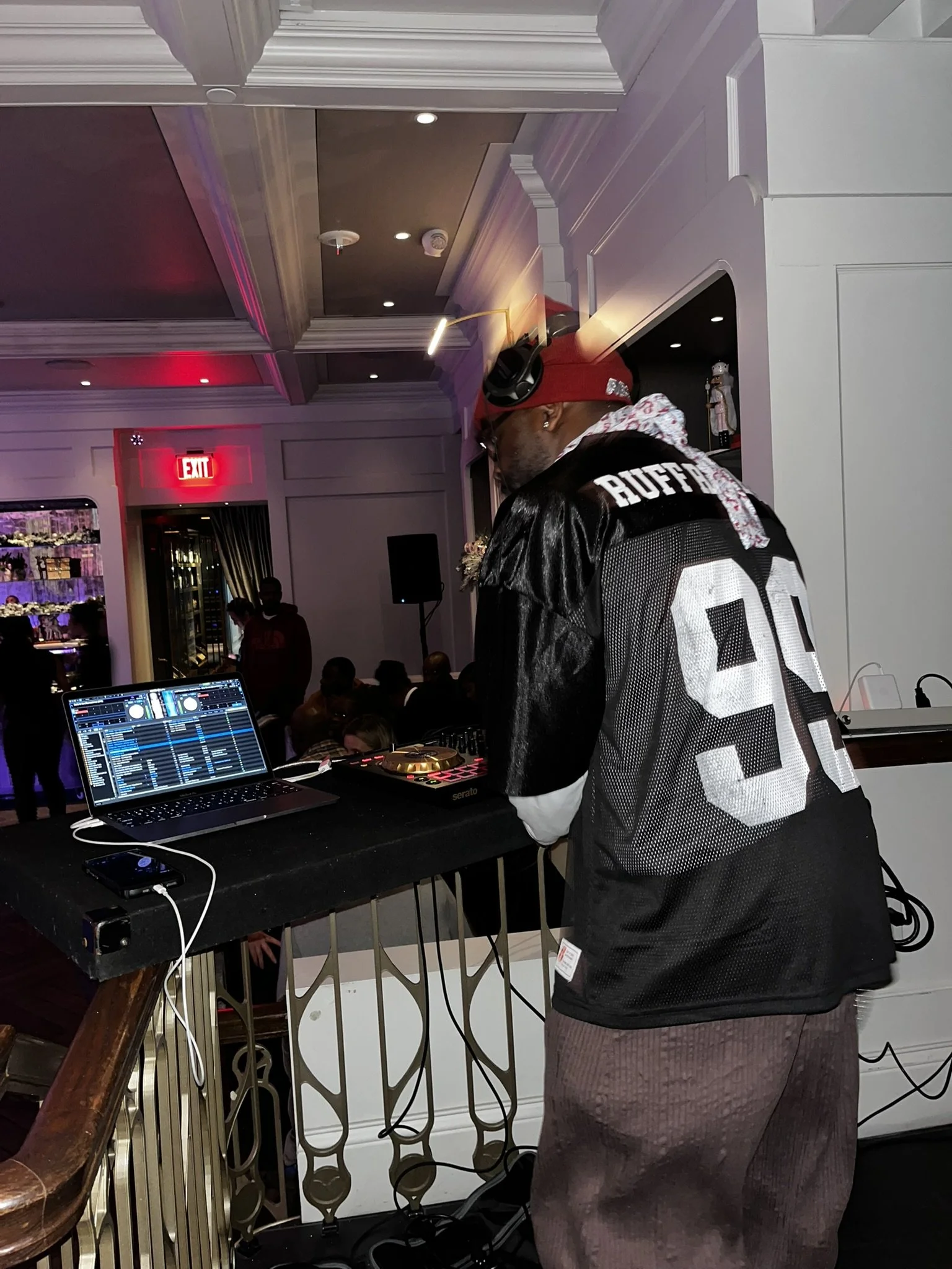 A DJ in a black football jersey, red cap, and headphones performing at a party, with a DJ mixer and laptop on a table, and people in the background in a decorated indoor venue.