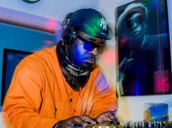 DJ wearing headphones, sunglasses, and a dark cap, working on a mixing console in a neon-lit studio.