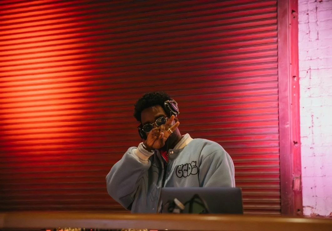 A person with curly hair wearing sunglasses and headphones, making a peace sign with their hand, standing in front of a red metal shutter with pink lighting on a white brick wall behind.