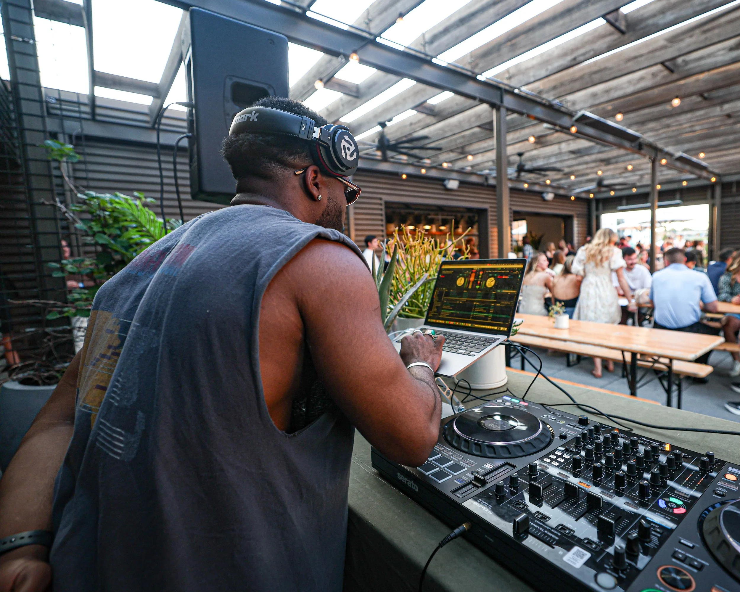 A DJ wearing headphones and a sleeveless shirt sets up music equipment with a crowd of people enjoying themselves in the background at an outdoor event.