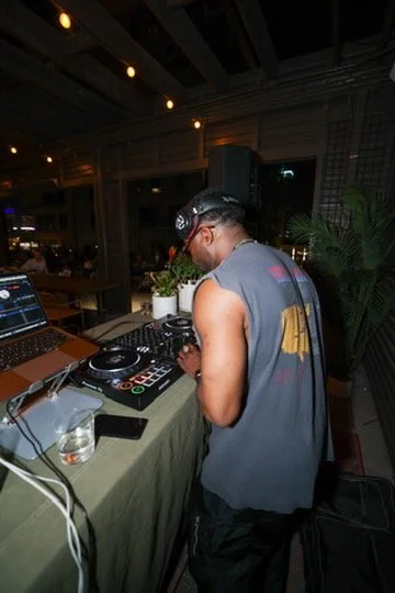 DJ wearing headphones and sleeveless shirt, using DJ equipment at a party or event.