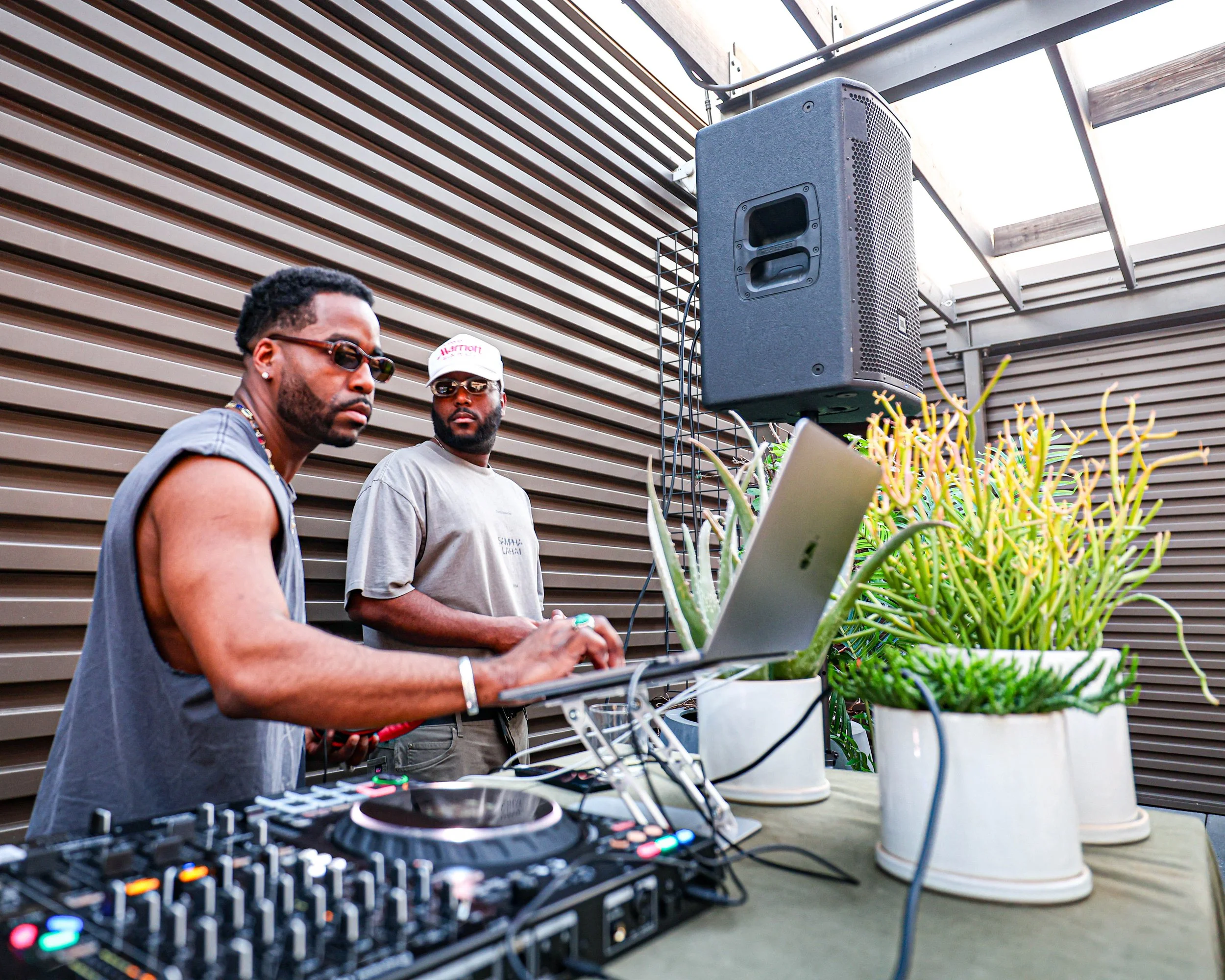 Two men at an outdoor DJ setup with a laptop, mixer, large speaker, and potted plants on a table against a brown louvered wall.